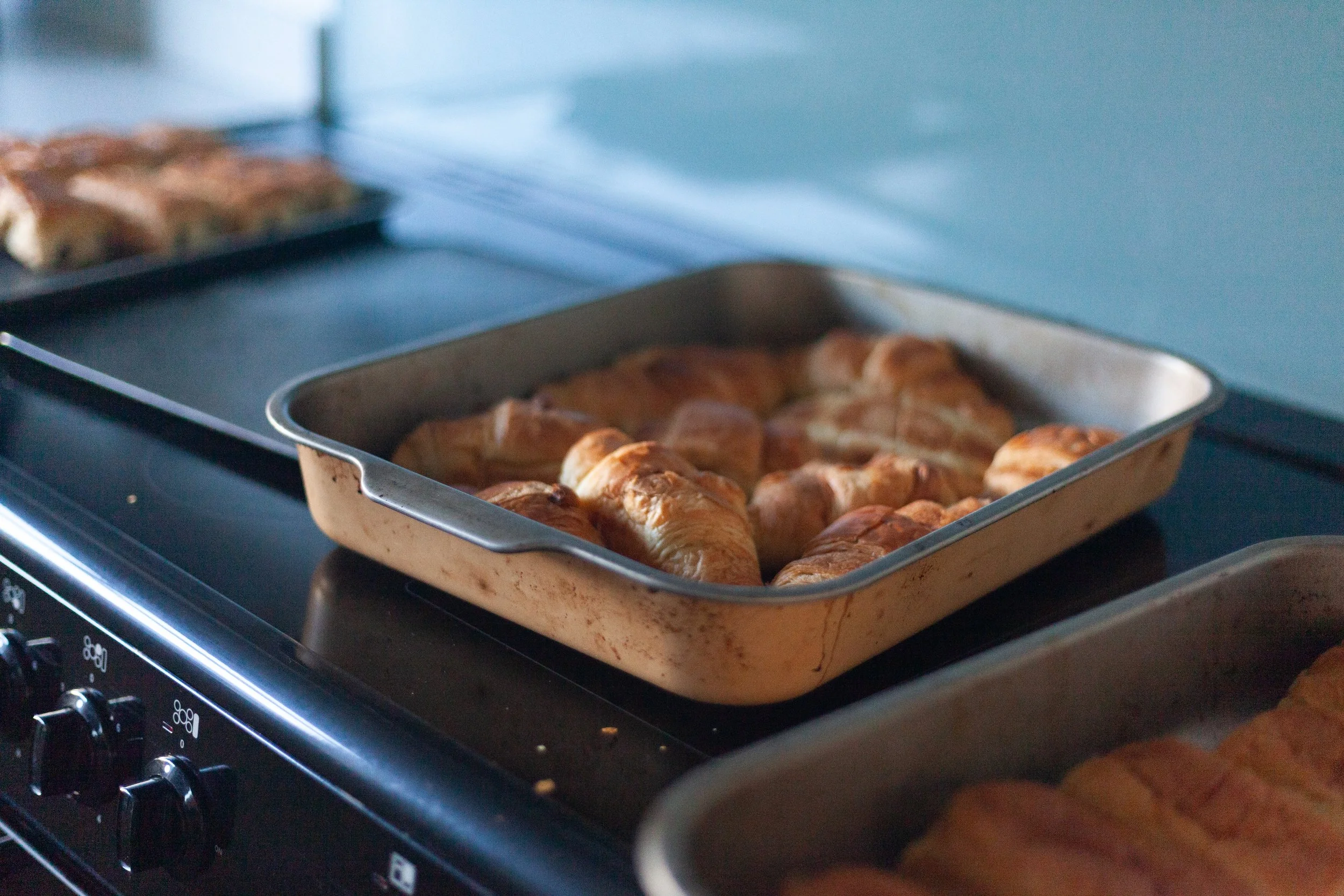 Baked croissants in a metal baking tray on a stovetop with additional pastries on a tray in the background.