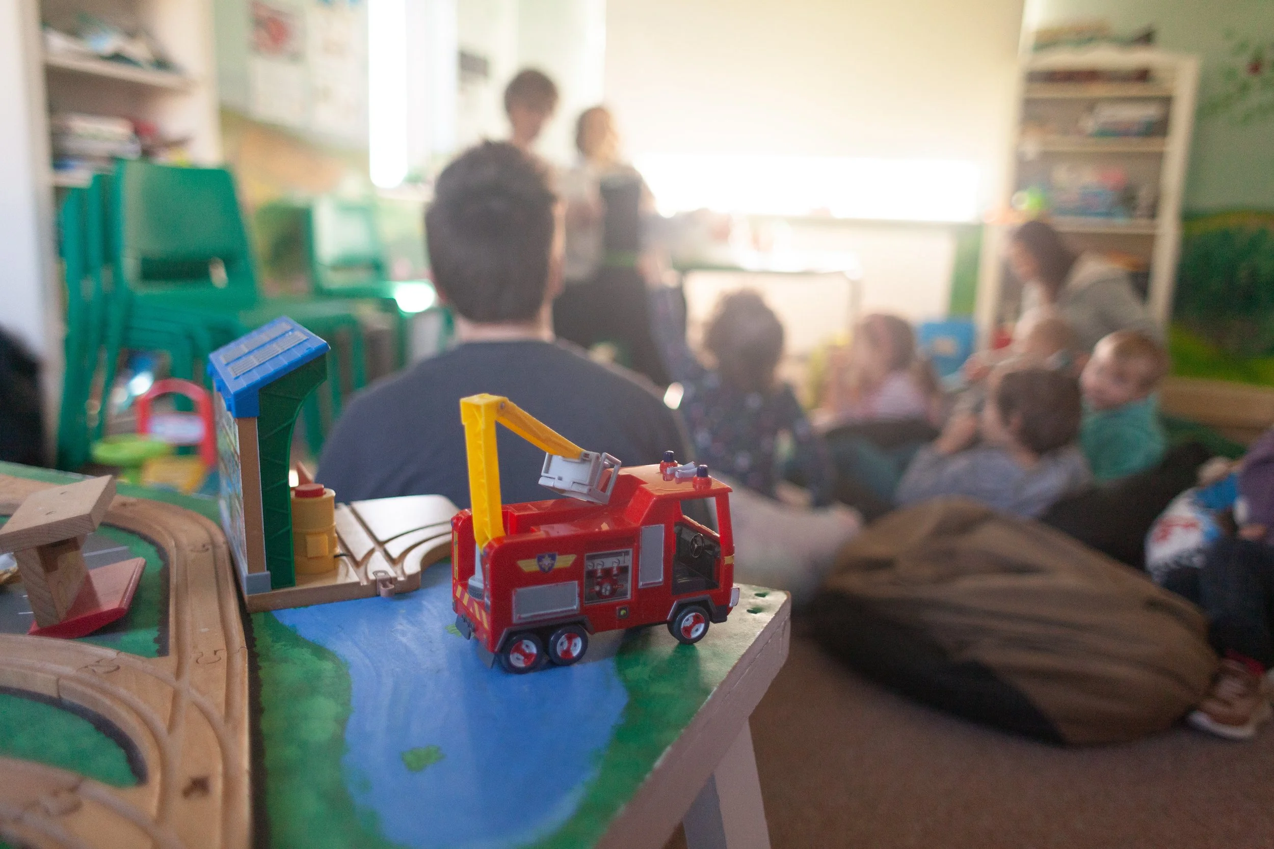 Toy fire truck on a play table in the foreground with a classroom scene in the background, including children and adults sitting and standing near a window.