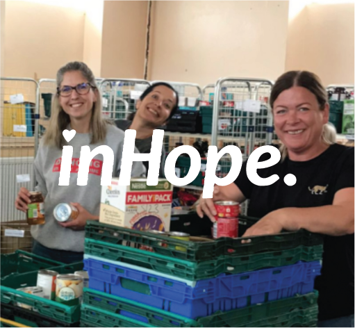 Three women smile behind stacks of canned goods and a boxed food item at a food bank or pantry.