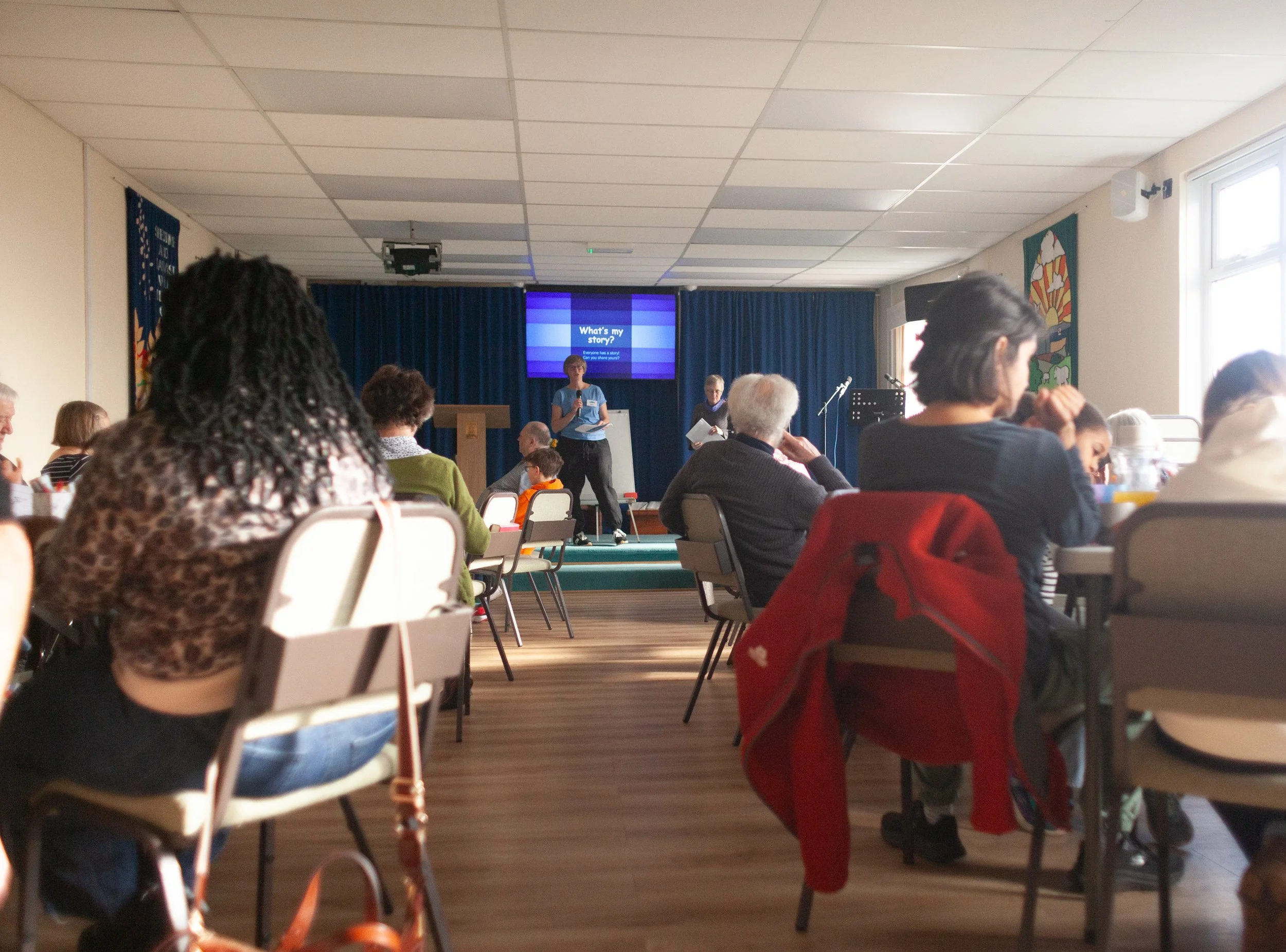 An indoor community event with people seated at tables, a stage with three speakers, and a screen displaying "What's my story?"