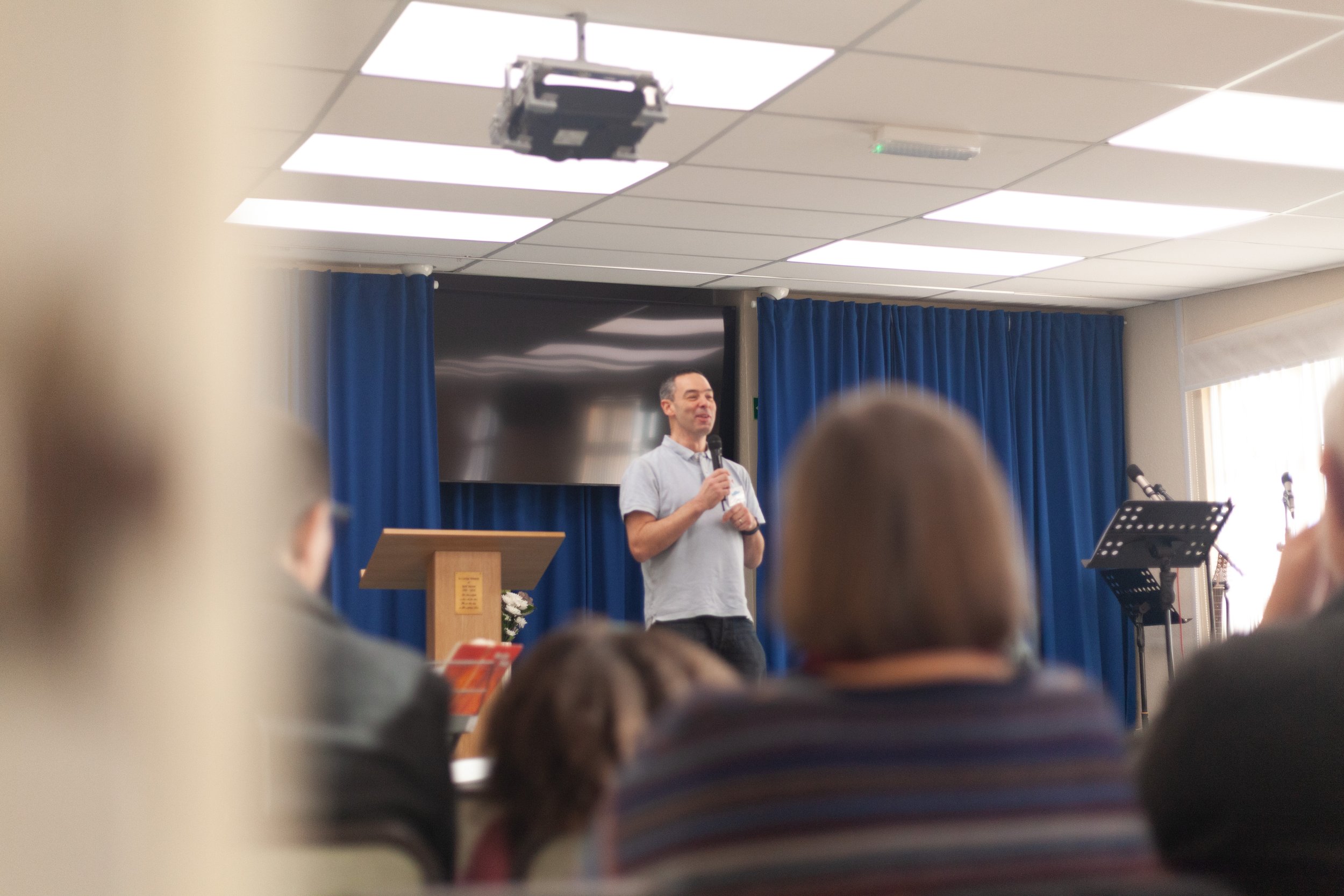 A man holding a microphone and speaking to an audience in a room with blue curtains and a large screen behind him, with several people seated and listening.
