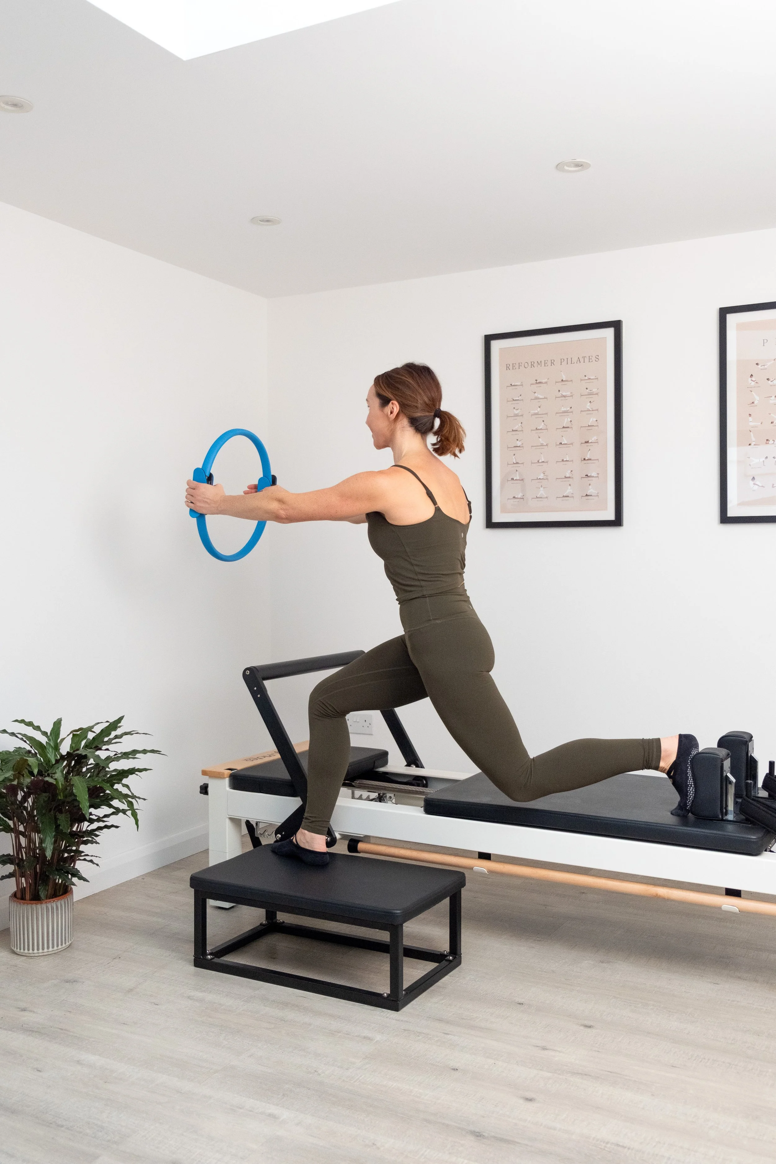 A woman in a dark green athletic outfit is doing Pilates using a reformer machine with a blue circle accessory, in a bright, minimalistic room with framed artwork on the wall and a potted plant in the corner.