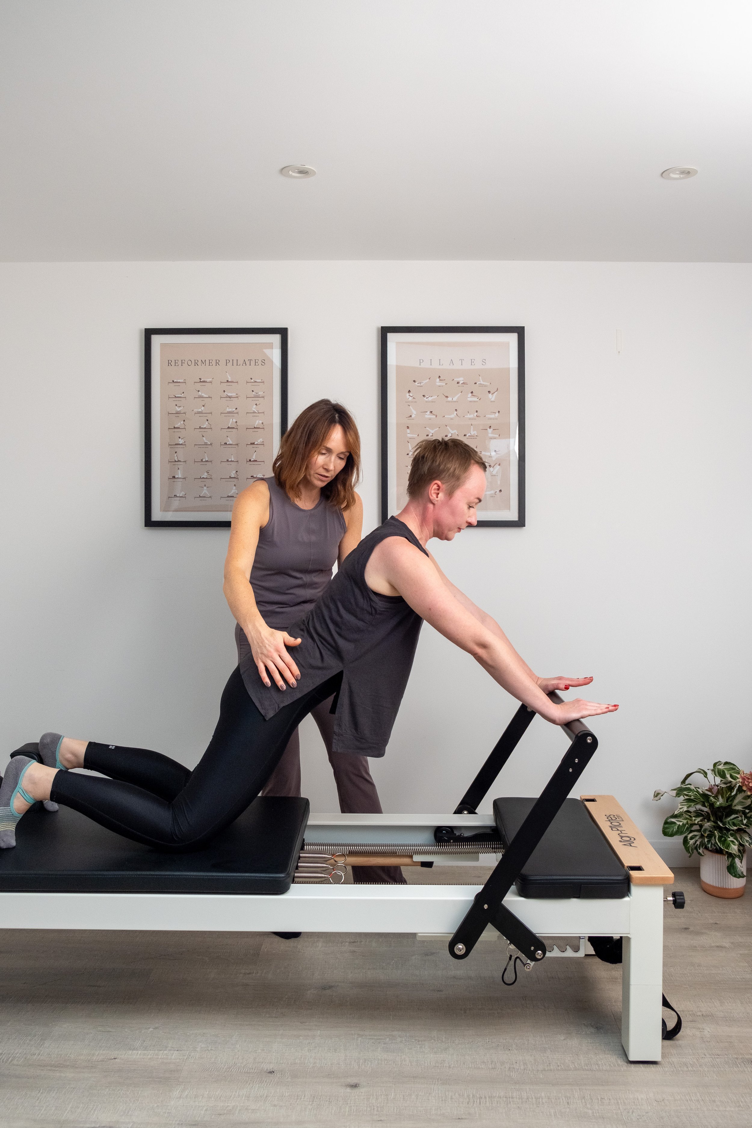 A woman is receiving a Pilates exercise session from an instructor on a Pilates reformer machine in a studio with framed Pilates posters on the wall and a potted plant in the corner.