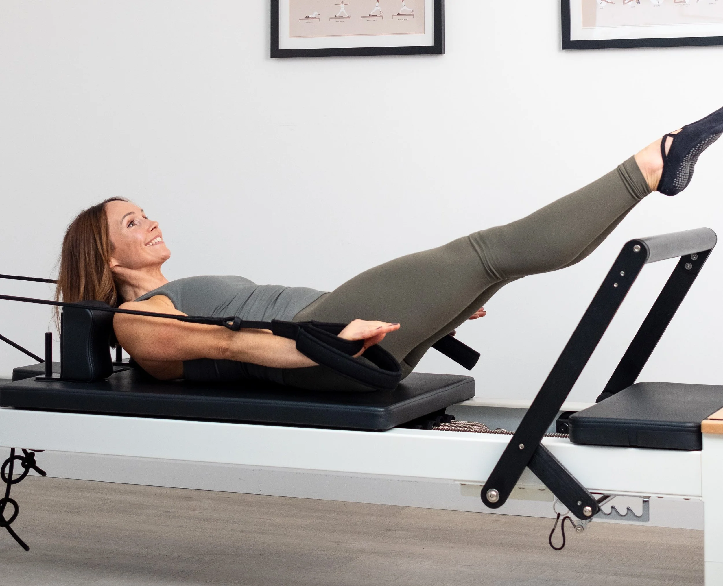 A woman doing a Pilates exercise on a reformer machine, smiling and lying on her back with her legs raised.
