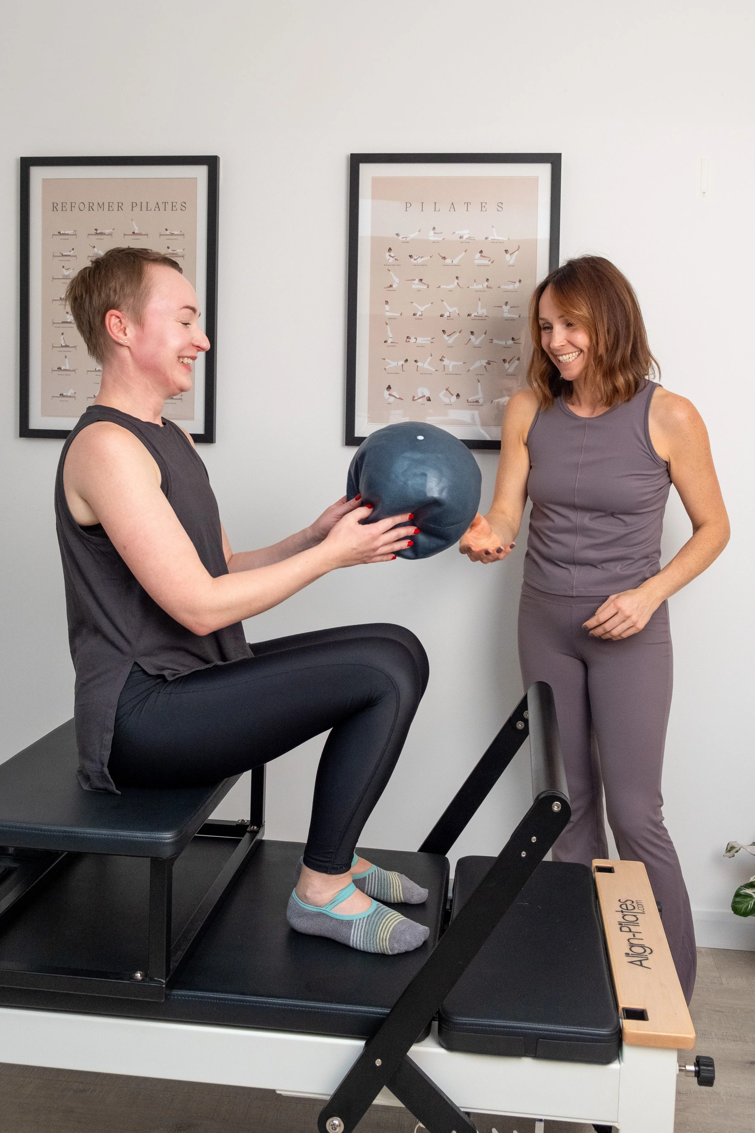 A woman receiving a blue exercise ball from a woman in a Pilates studio, with Pilates posters on the wall behind them.