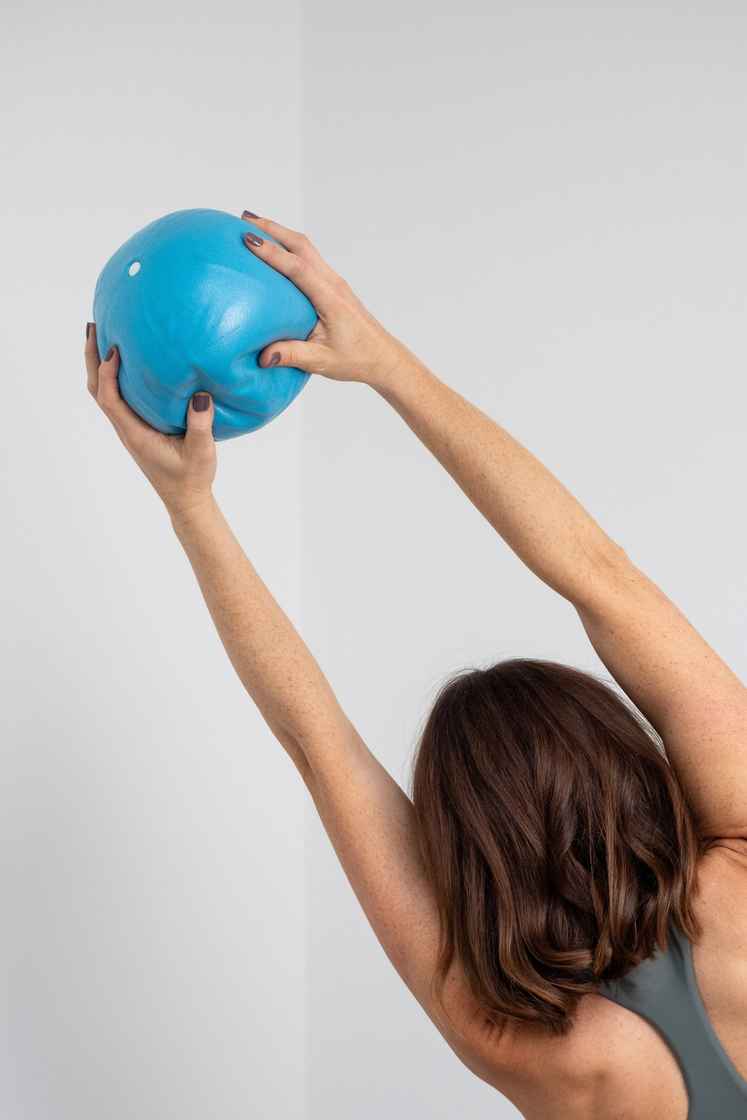 A woman with short brown hair stretching her arms overhead, holding a blue exercise ball in a gym or studio with plain white walls.