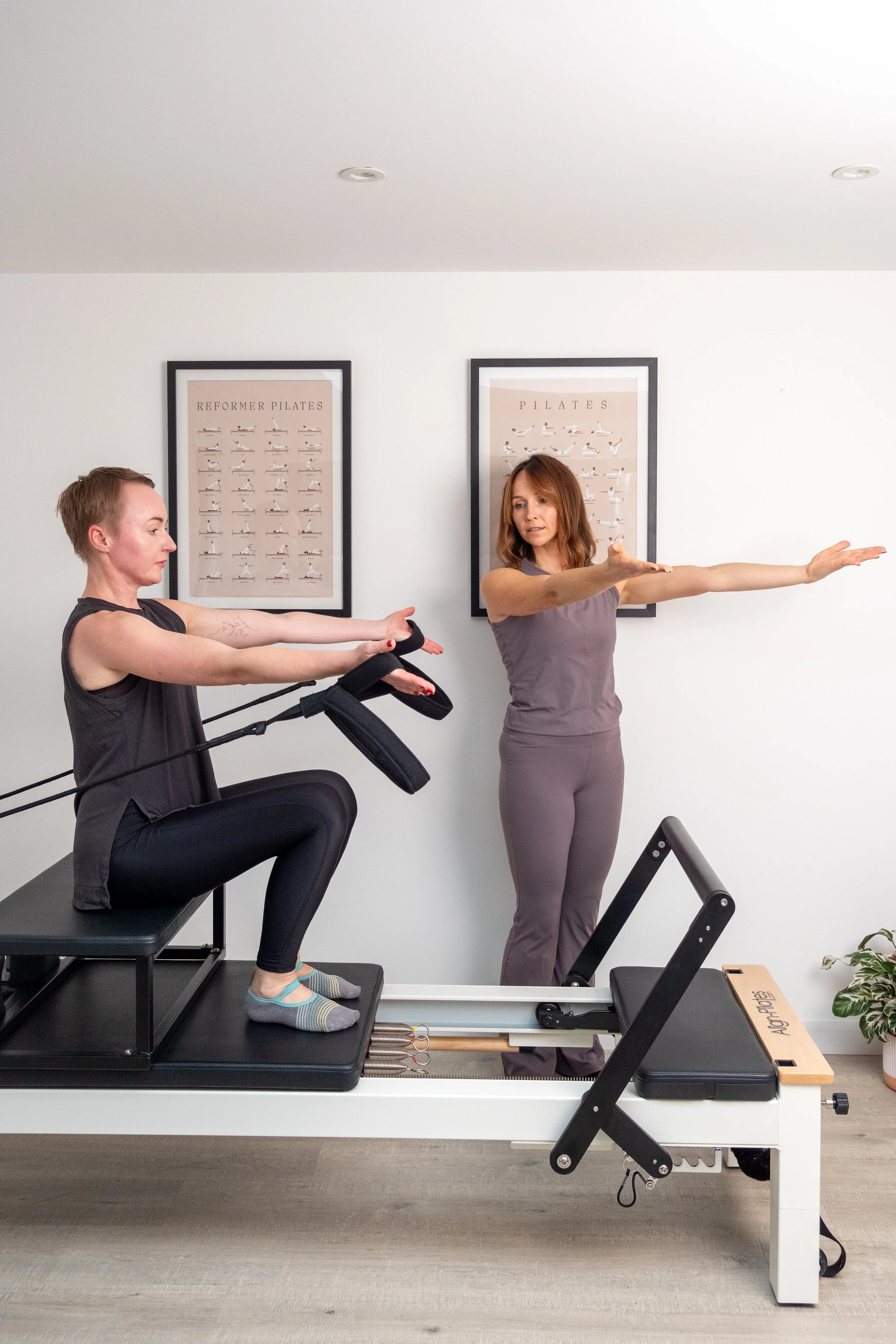 Two women practicing Pilates with reformer equipment in a studio, one sitting on the reformer and the other standing, both extending their arms in a stretching exercise.
