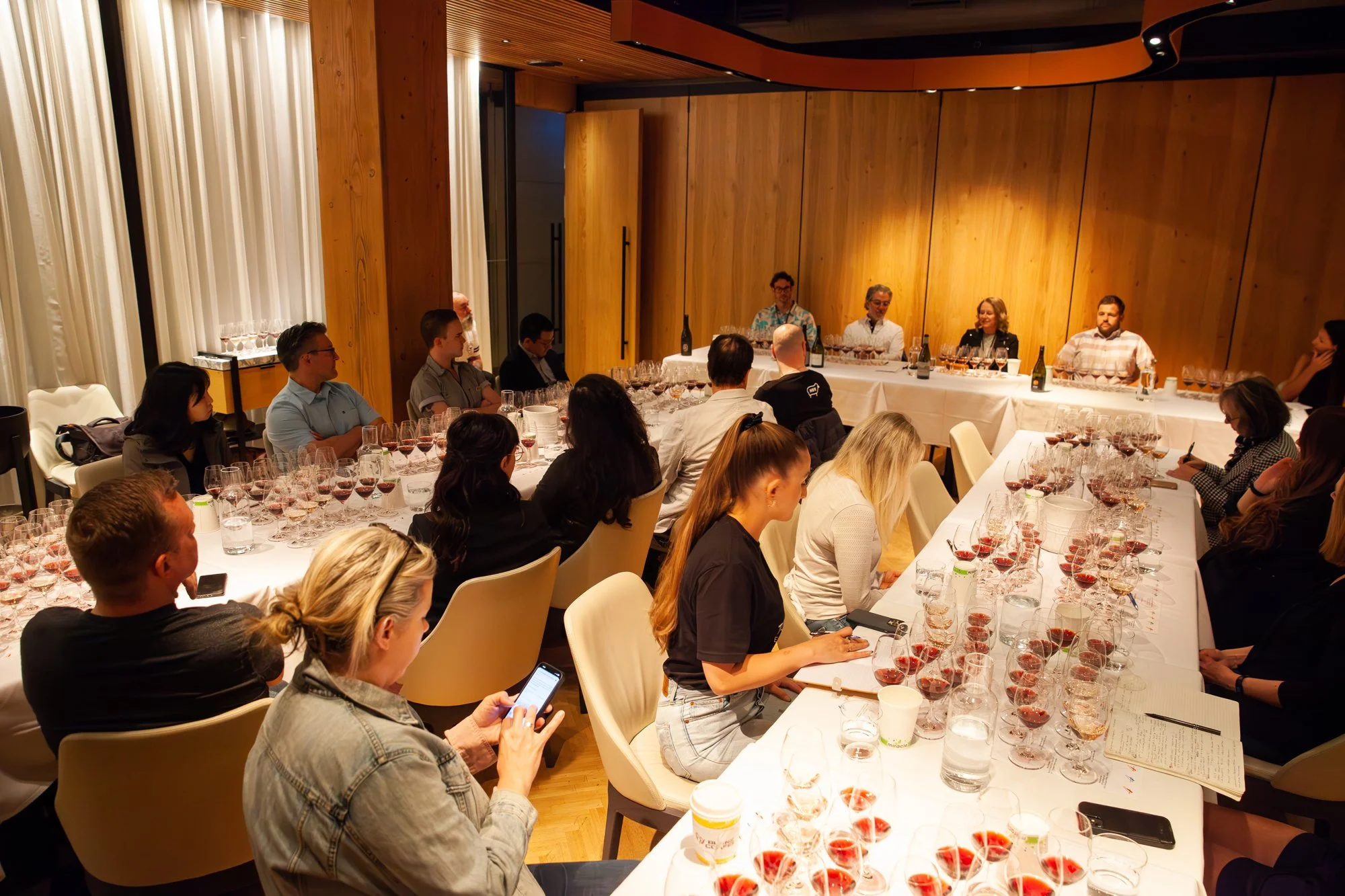 People attending a wine tasting event seated around tables with glasses of red wine, with some taking notes and others listening to the speakers at the front of the room at Top Drop Canada