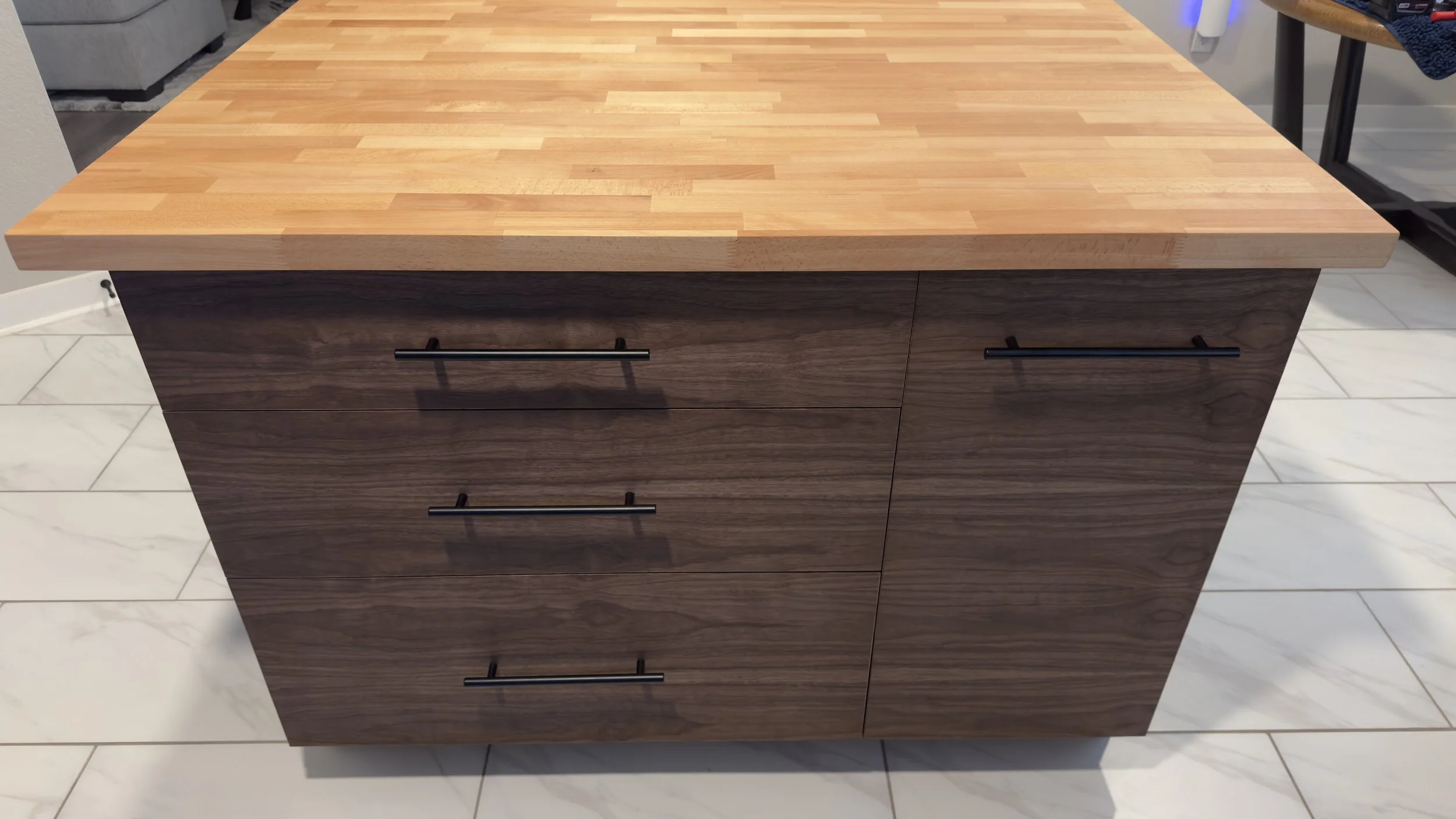 A wooden kitchen island with a light-colored butcher block countertop and dark wood drawers with black handles, situated on a tiled floor.