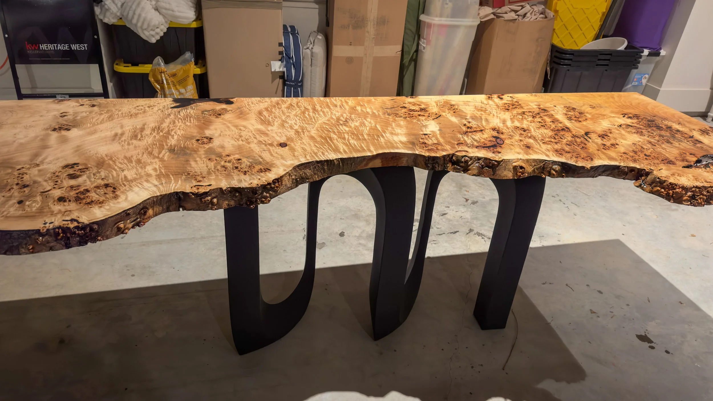 A live edge wood table with black metal legs in a workshop. The table has a natural, irregular edge and a polished, richly grained surface.