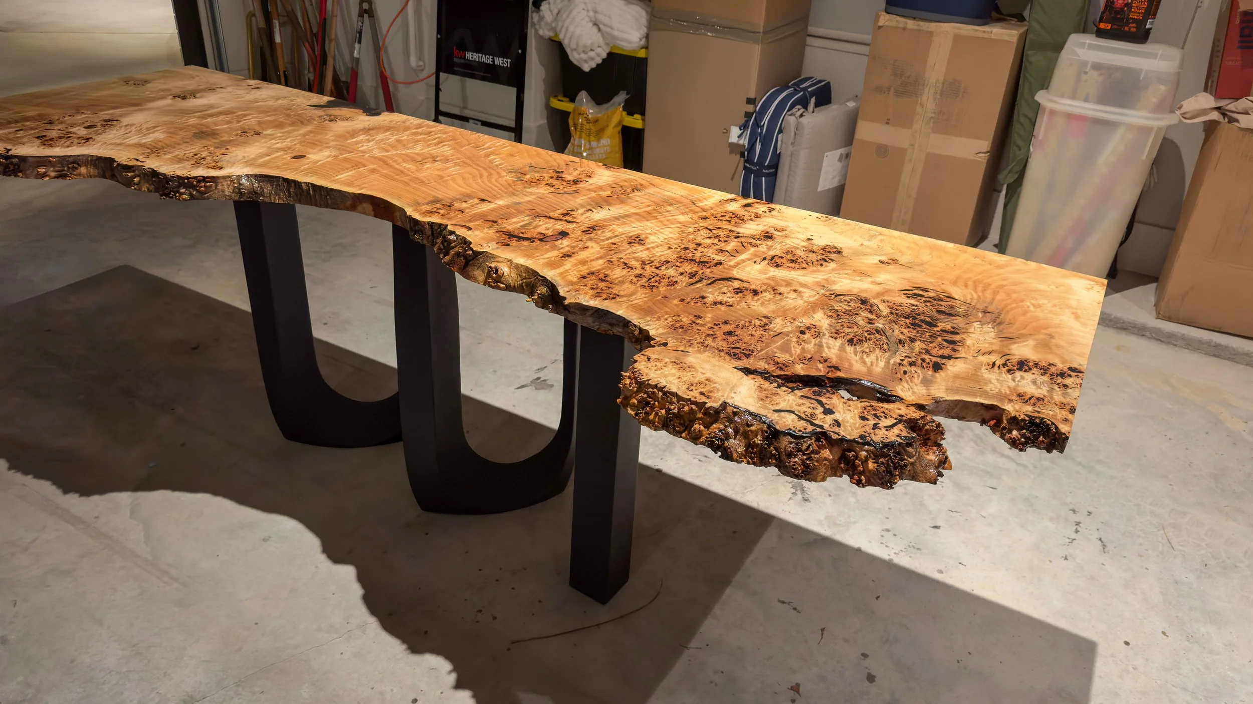 A live edge wooden table with a natural, irregular edge and a highly figured burl pattern, supported by black U-shaped metal legs, in a garage setting.