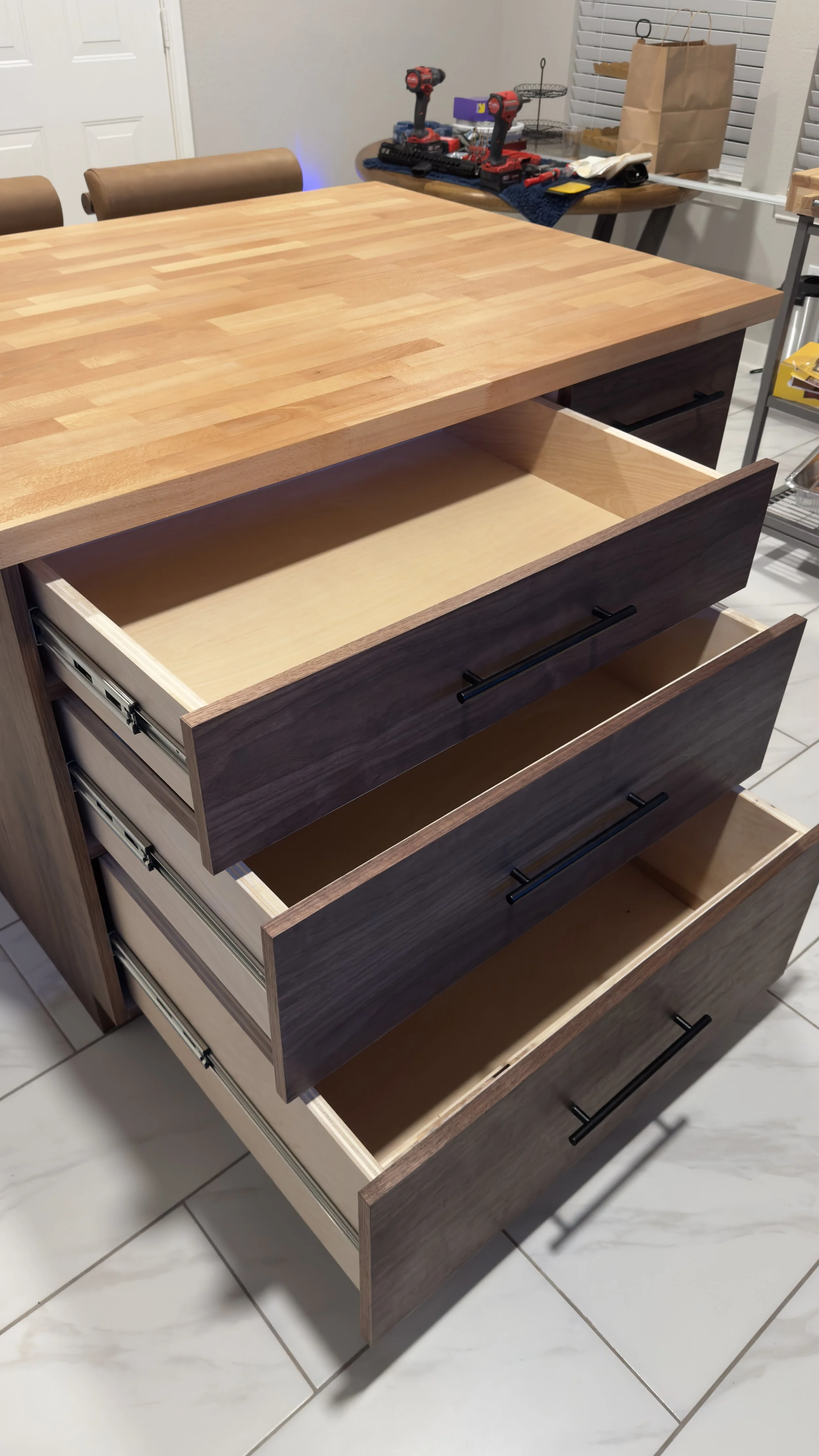 A wooden kitchen island with three open drawers, showing the interior of the drawers. The island has a butcher block countertop, and the background includes a table with power drills, a paper bag, and miscellaneous items.
