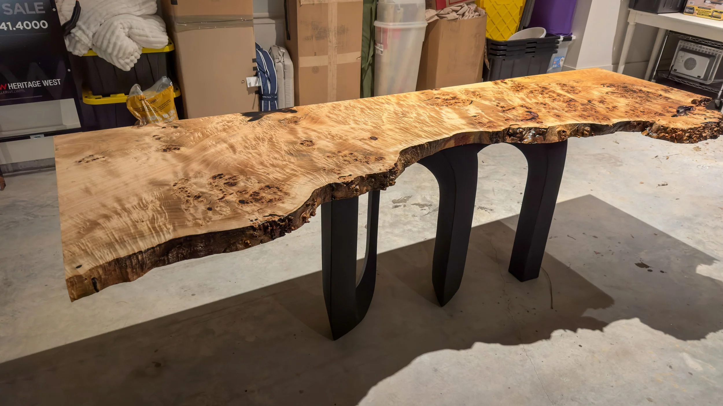 A live edge wooden table with a natural, irregular edge and a glossy finish, supported by two black curved legs, situated in a workshop or garage.