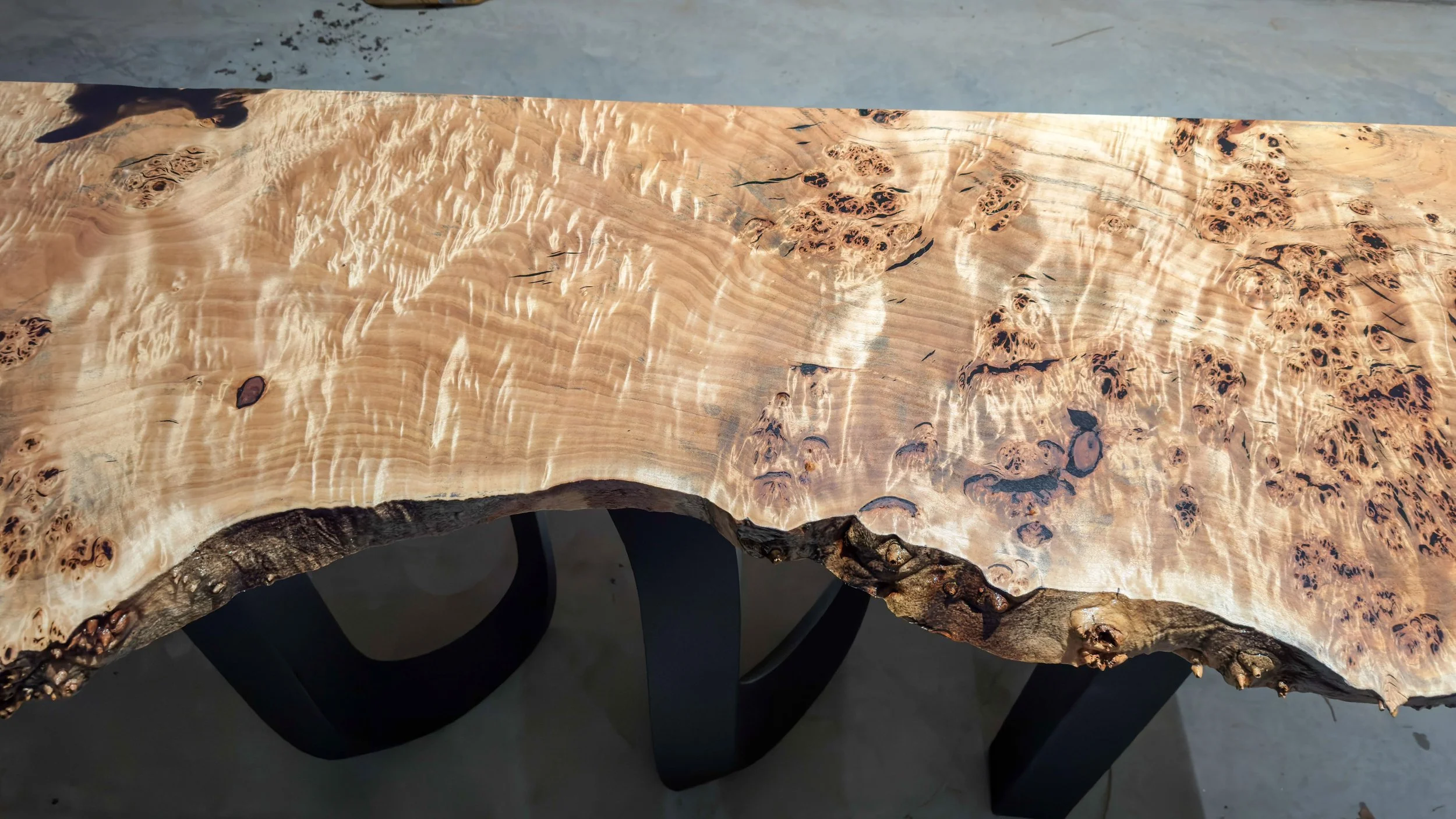 Close-up of a natural wood table with intricate grain patterns and a live edge, supported by a black metal base.