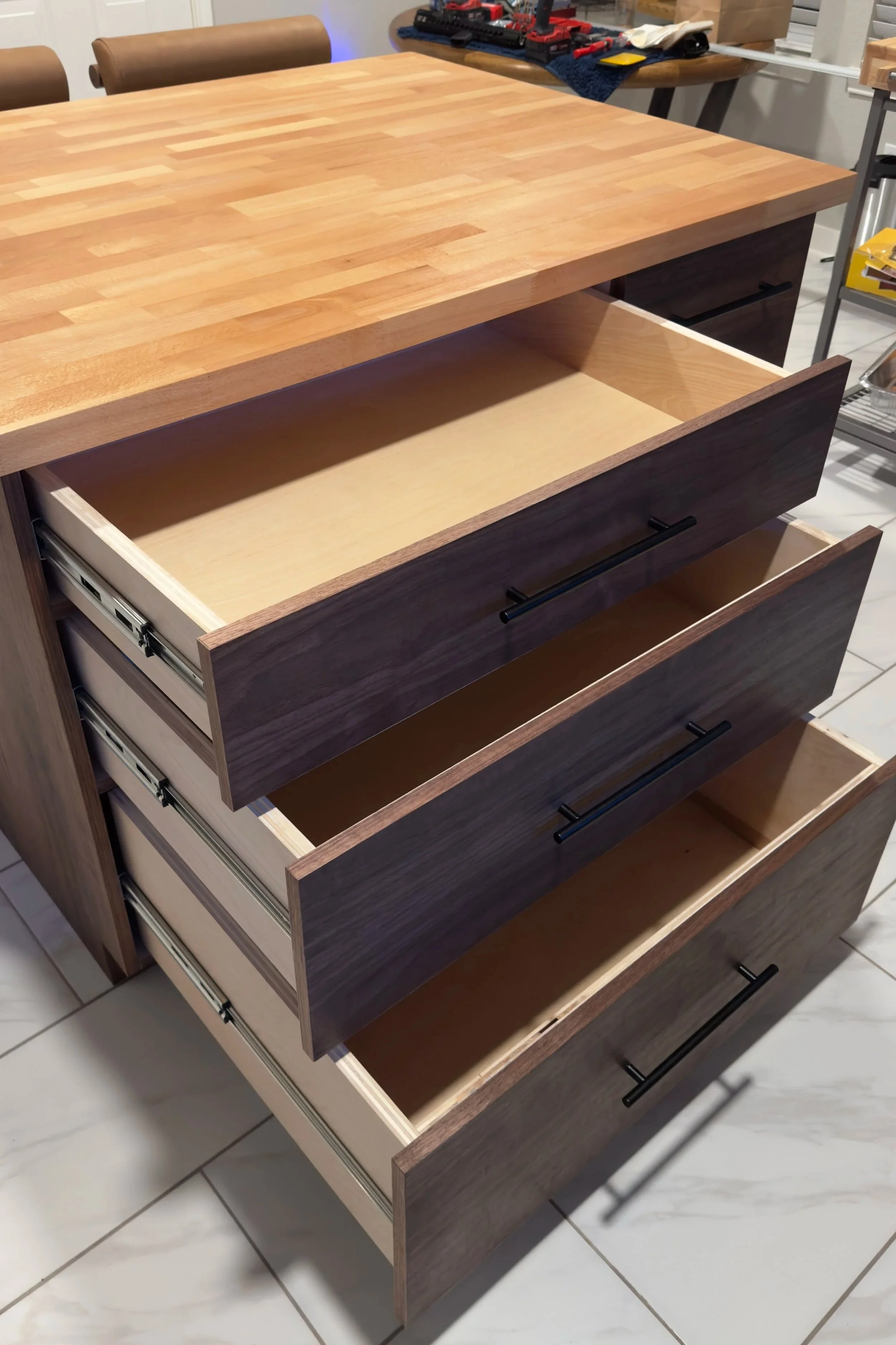 A wooden kitchen island with open drawers showing blank interiors, placed on a tiled floor. In the background, there are tools and other items on a workbench.