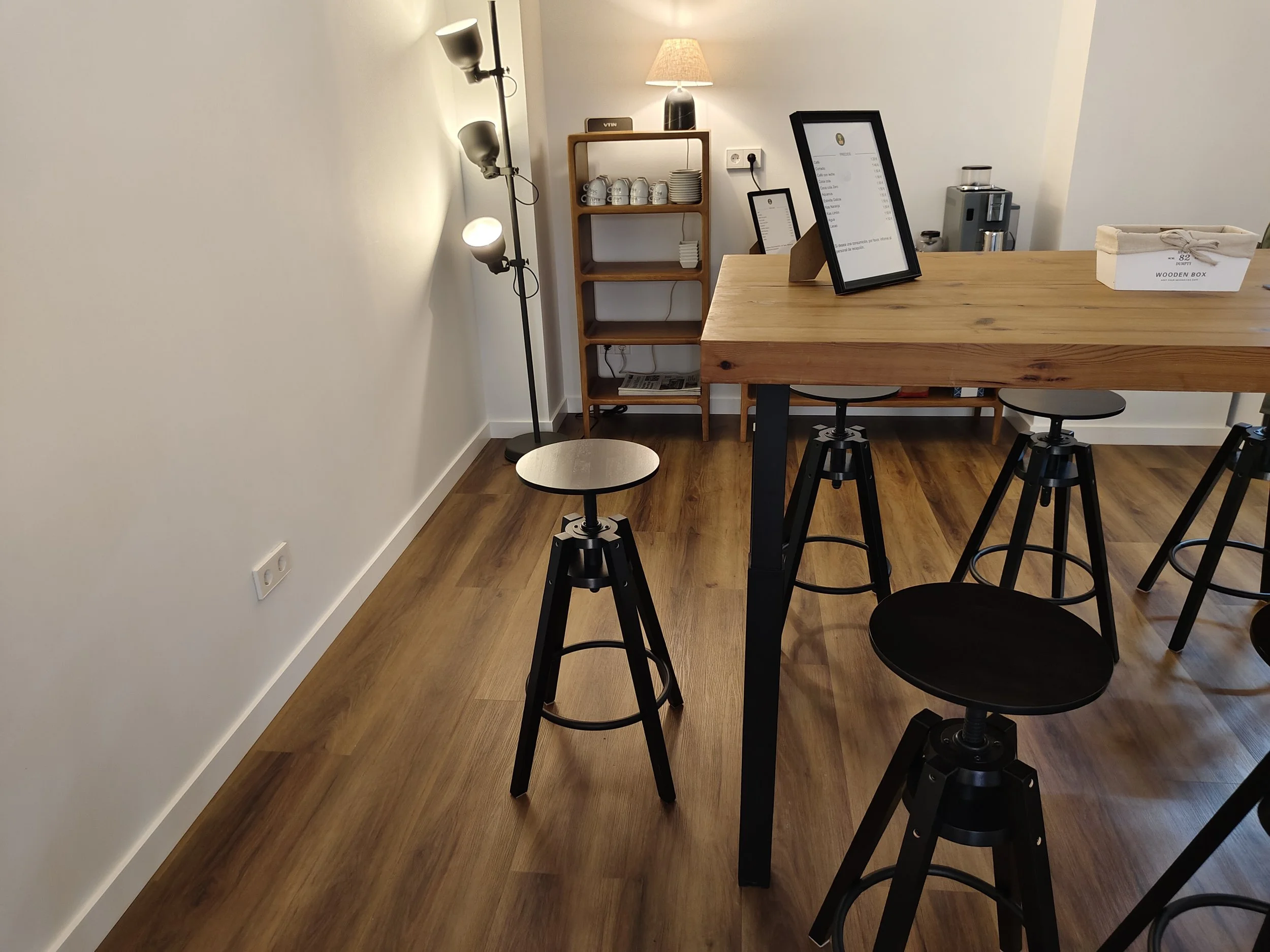 Interior de una cafetería moderna con barra de madera, taburetes negros, estanterías con tazas y platos, lámpara de mesa y luz de pie, y un ambiente ordenado y minimalista.