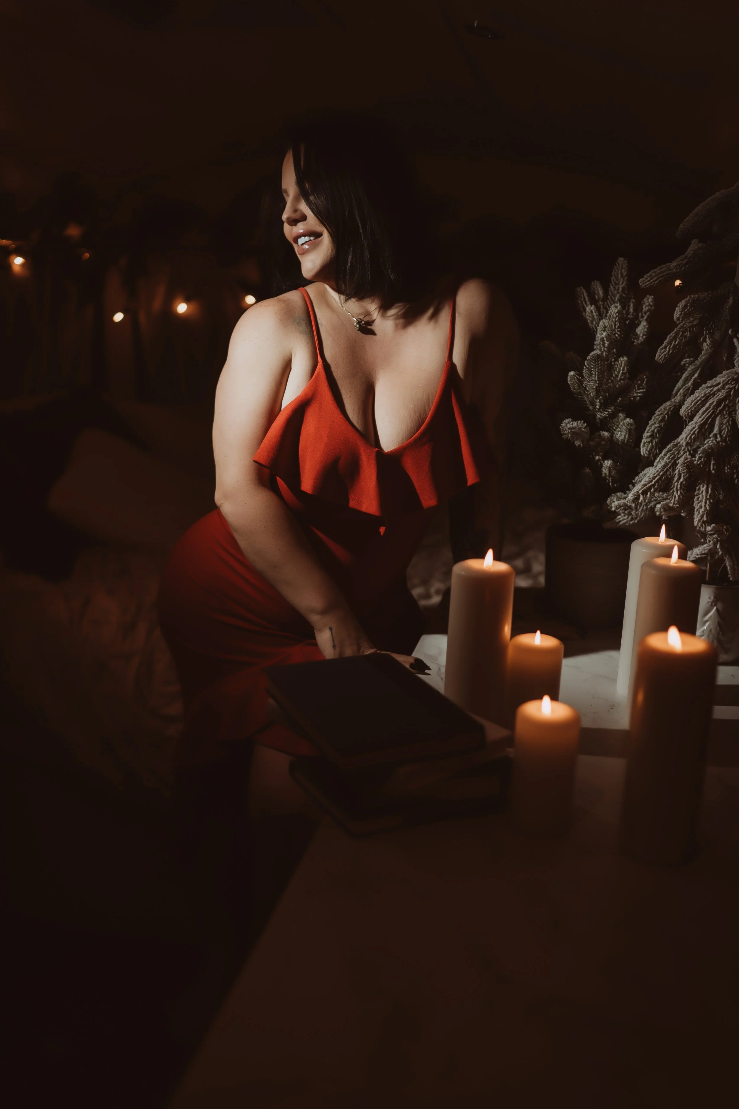 A woman in a red dress with ruffled details, smiling and looking to the side, surrounded by lit candles in a dimly lit room decorated for Christmas.