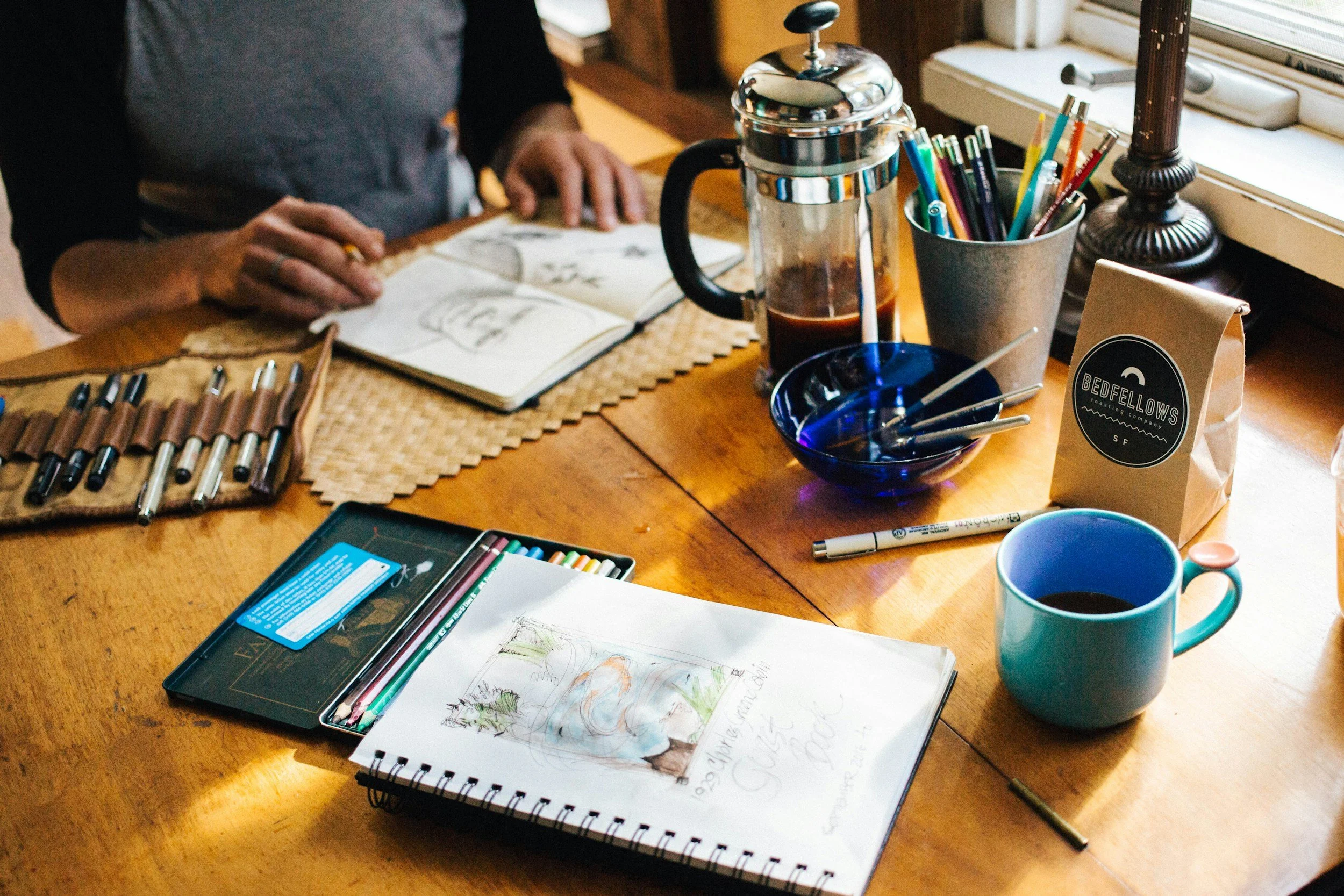 A cozy workspace with a wooden table filled with art supplies, a sketchbook with colorful drawings, pens, markers, a coffee cup, a French press with coffee, a bowl of paintbrushes, and a newspaper bag labeled "Bedfellows." Someone is drawing in an open sketchbook while working on art.