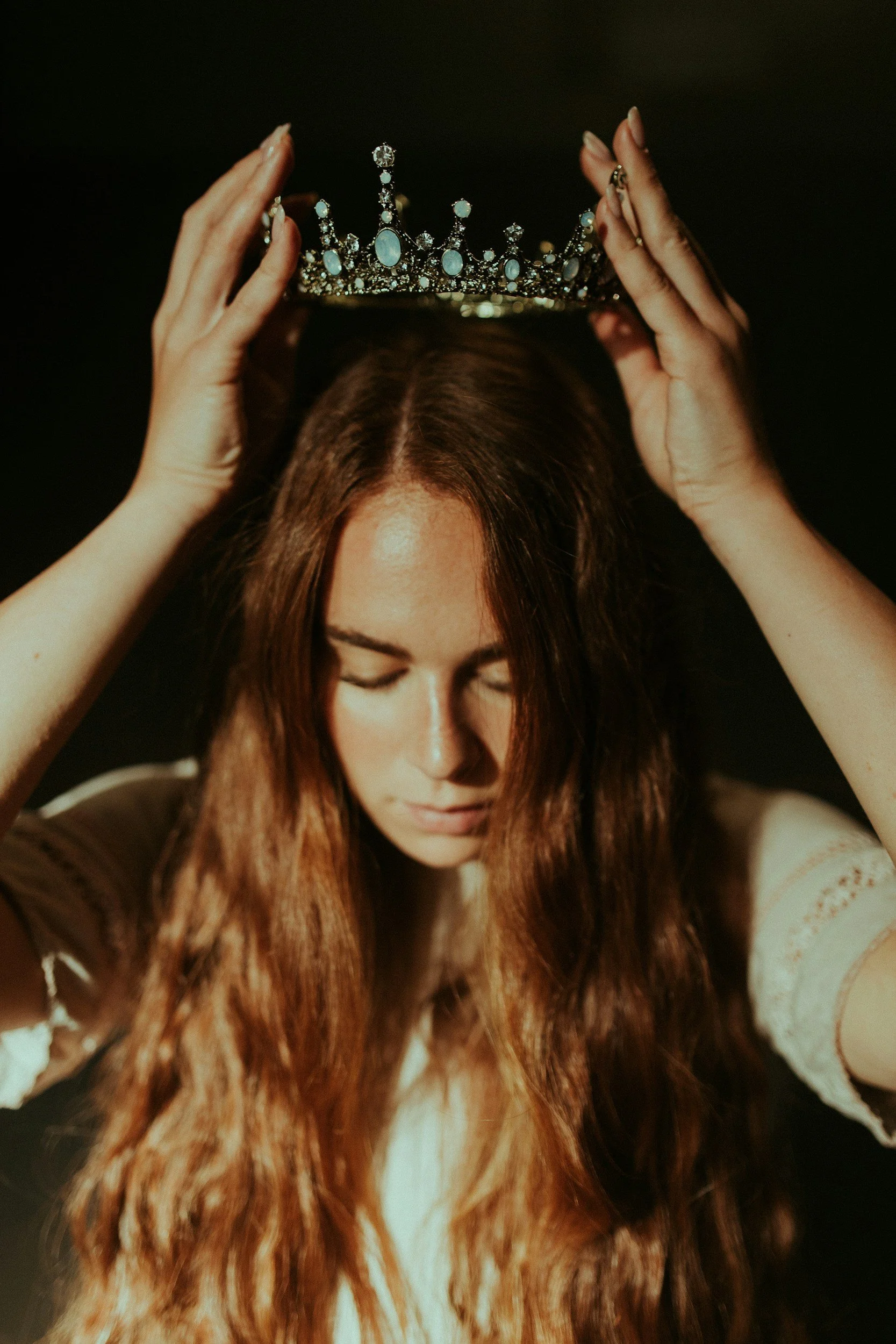 A woman with long red hair gently places a jeweled tiara on her head against a dark background, with her eyes closed.