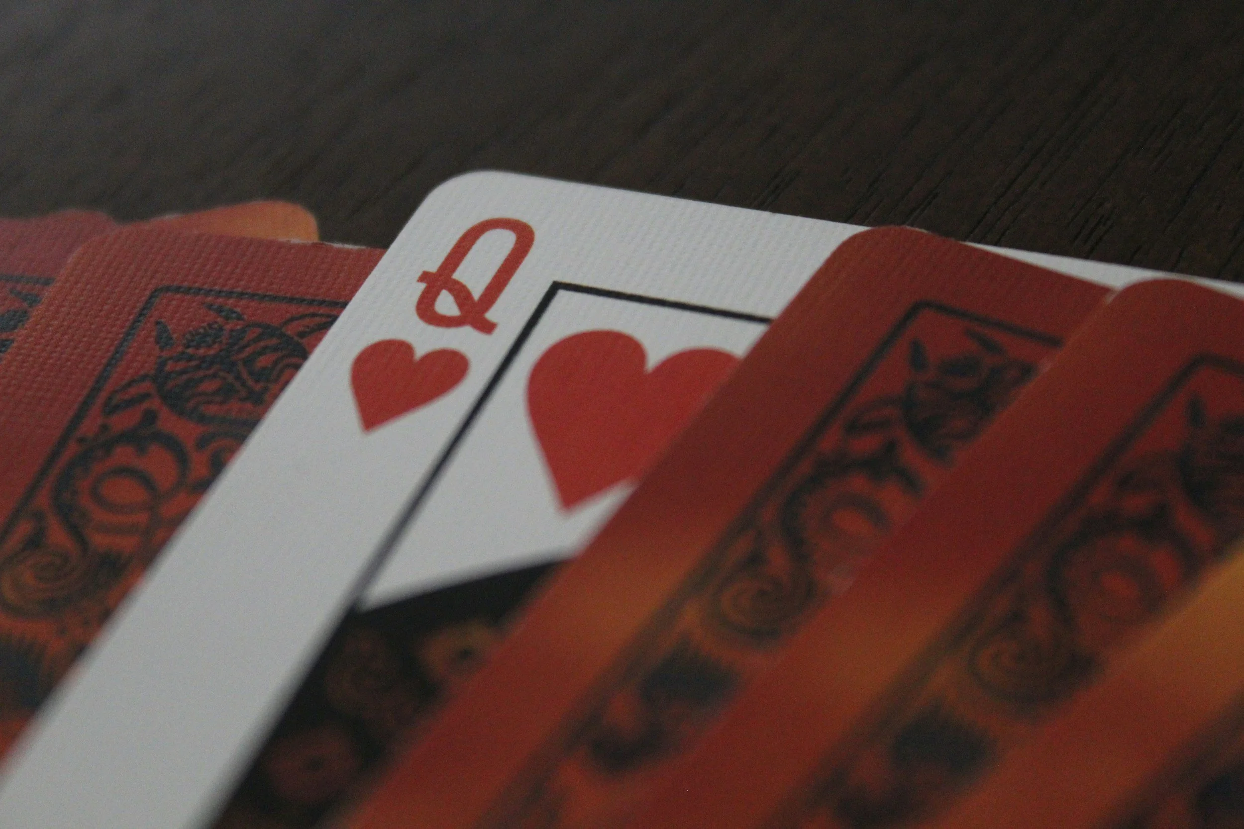 A close-up of a Queen of Hearts playing card with several face-down cards behind it, on a dark wooden surface.