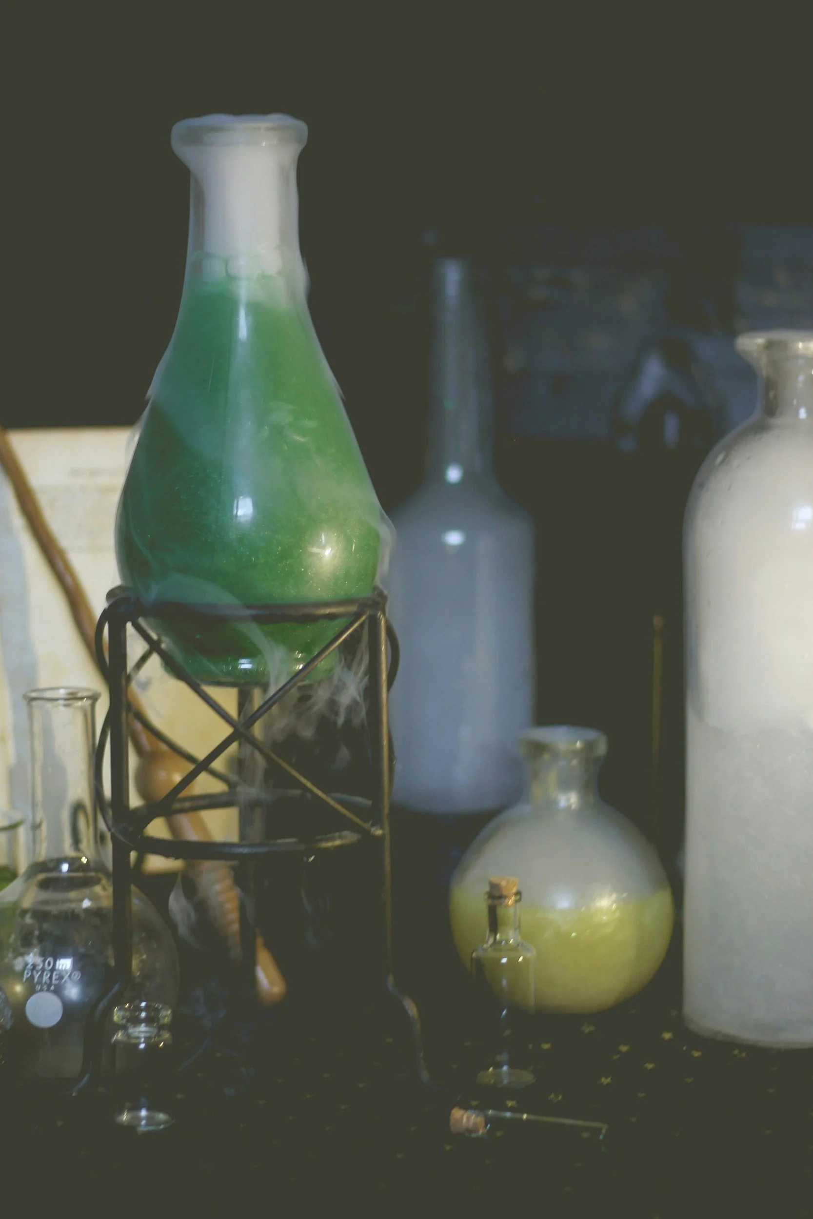 Several laboratory glass bottles and flasks containing colored liquids, one with green liquid emitting smoke, on a black surface.