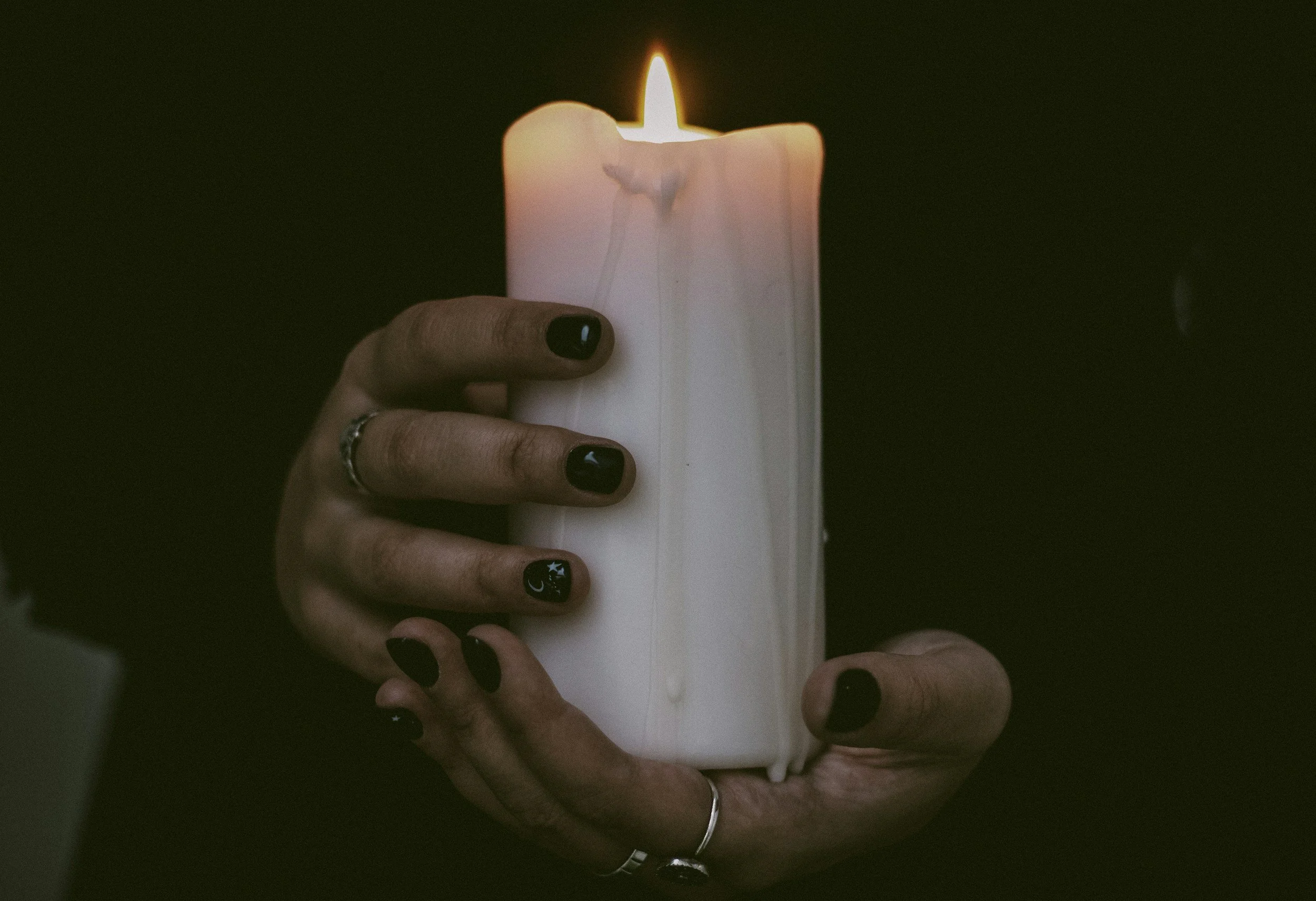 A hand with dark nail polish and rings holding a tall, burning white candle with wax melting down the sides.
