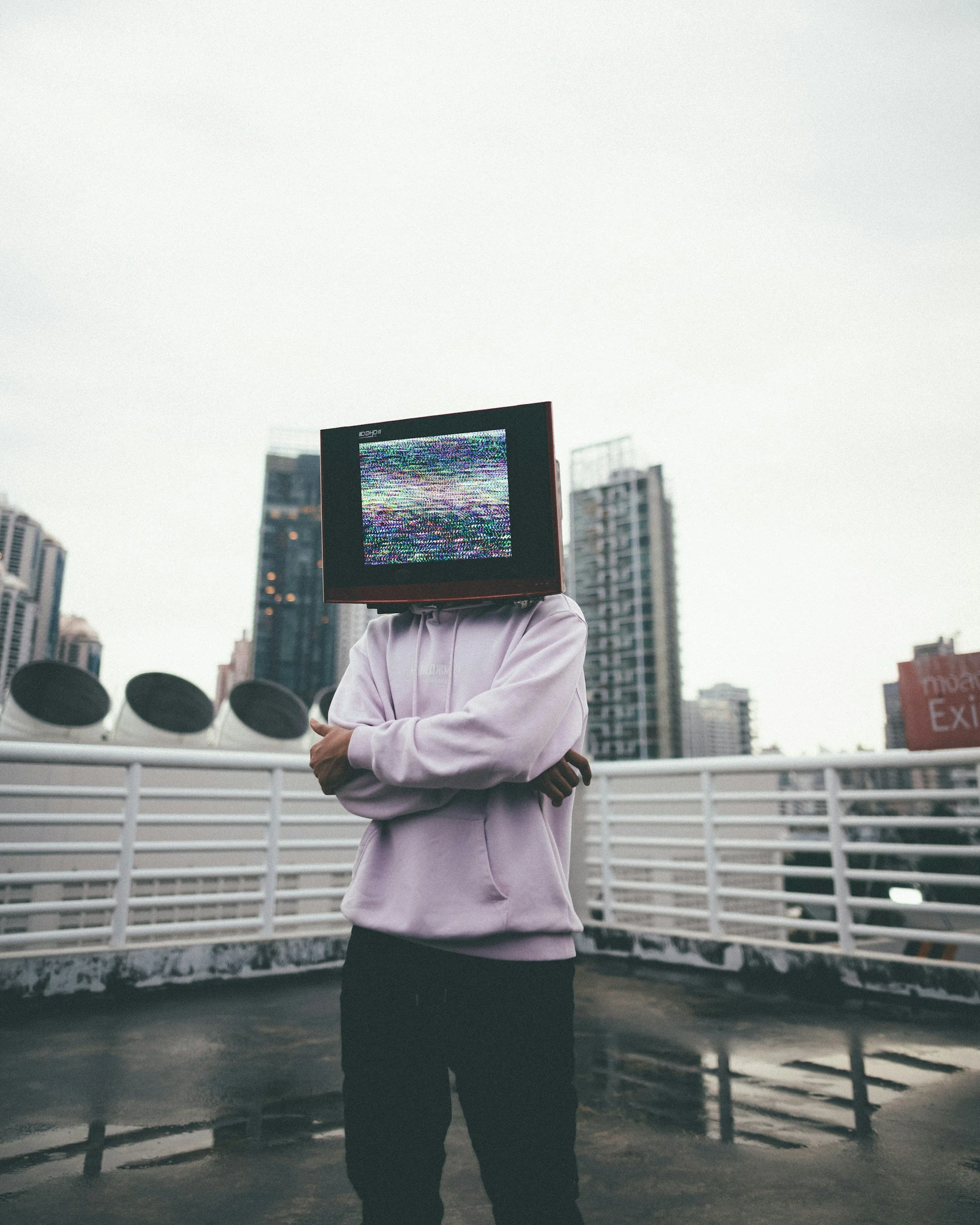 Person wearing a hoodie with a vintage television set on their head, standing on a rooftop with city buildings in the background.