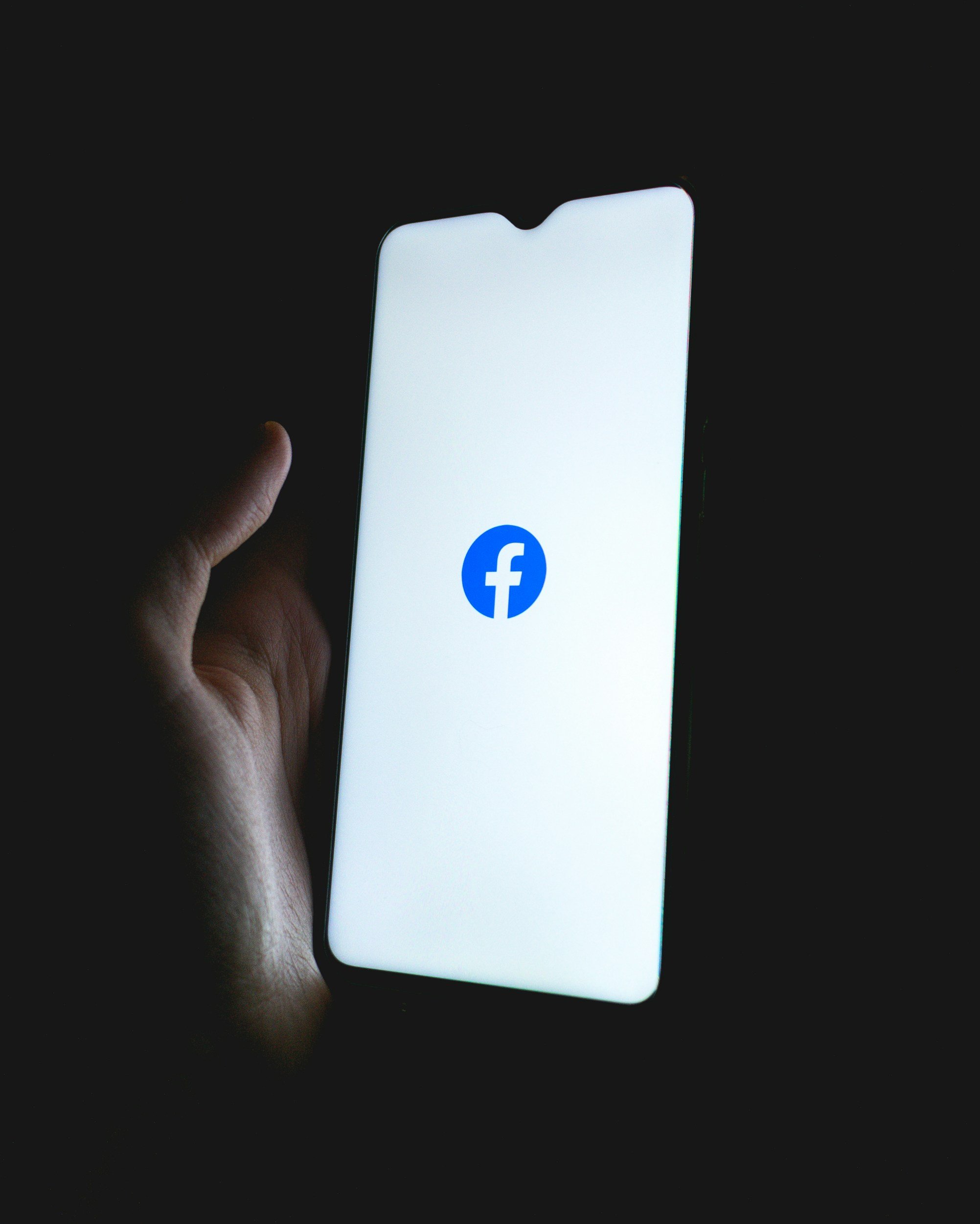 Hand holding a smartphone displaying the Facebook logo on a white background.