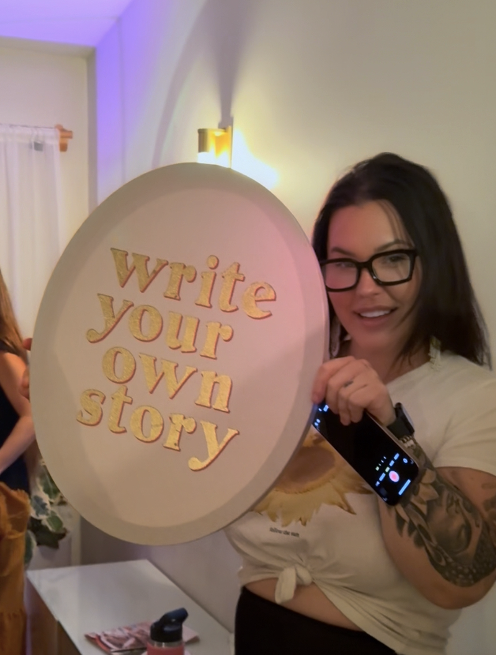 A woman with black hair and glasses holds a large balloon with the words 'write your own story' written on it. She is smiling and has tattoos on her arm, standing in a room with warm lighting.