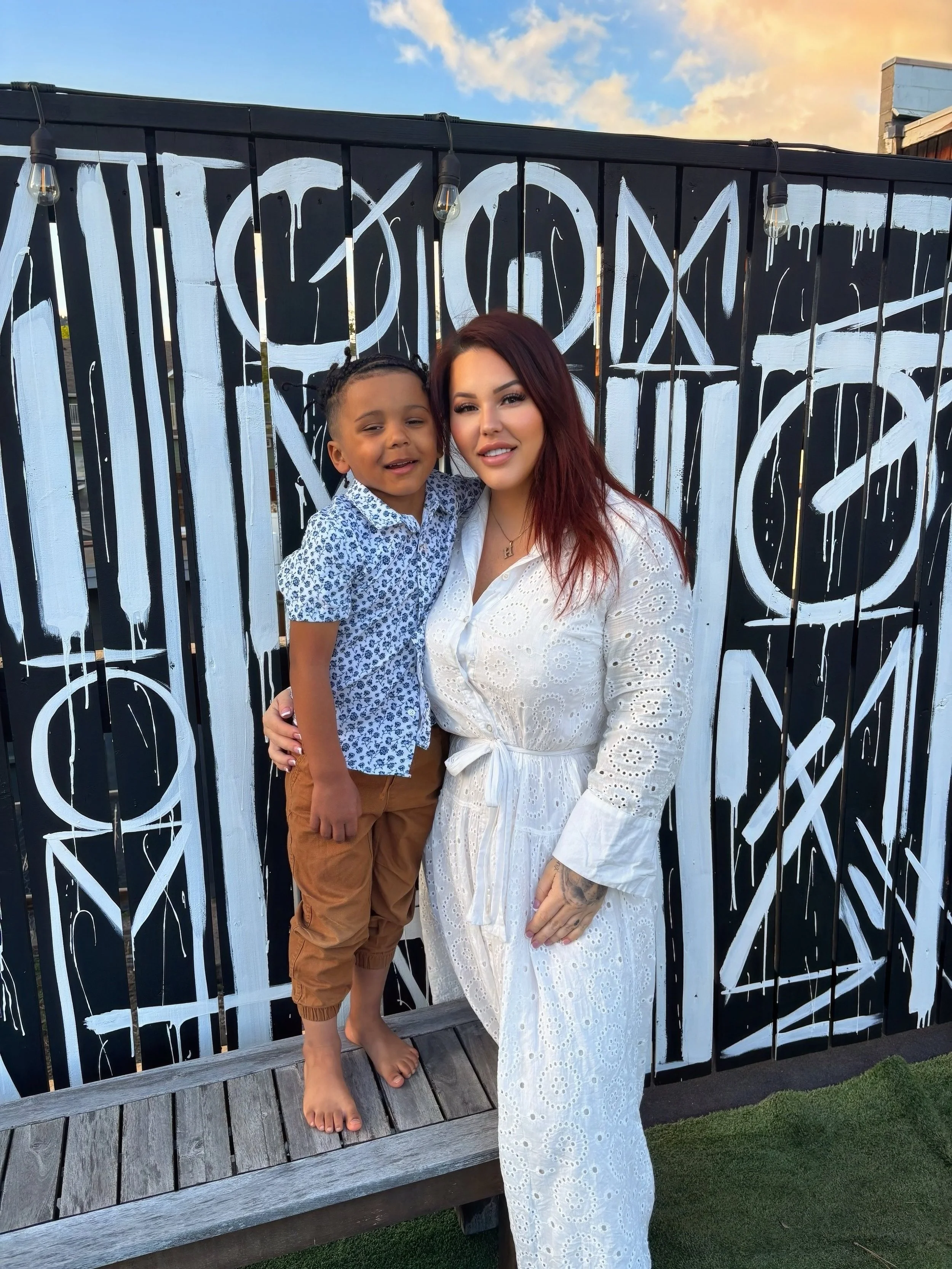 A woman with red hair wearing a white eyelet dress standing next to a young boy wearing a blue patterned shirt and tan pants, both smiling and posing in front of a black and white painted fence.