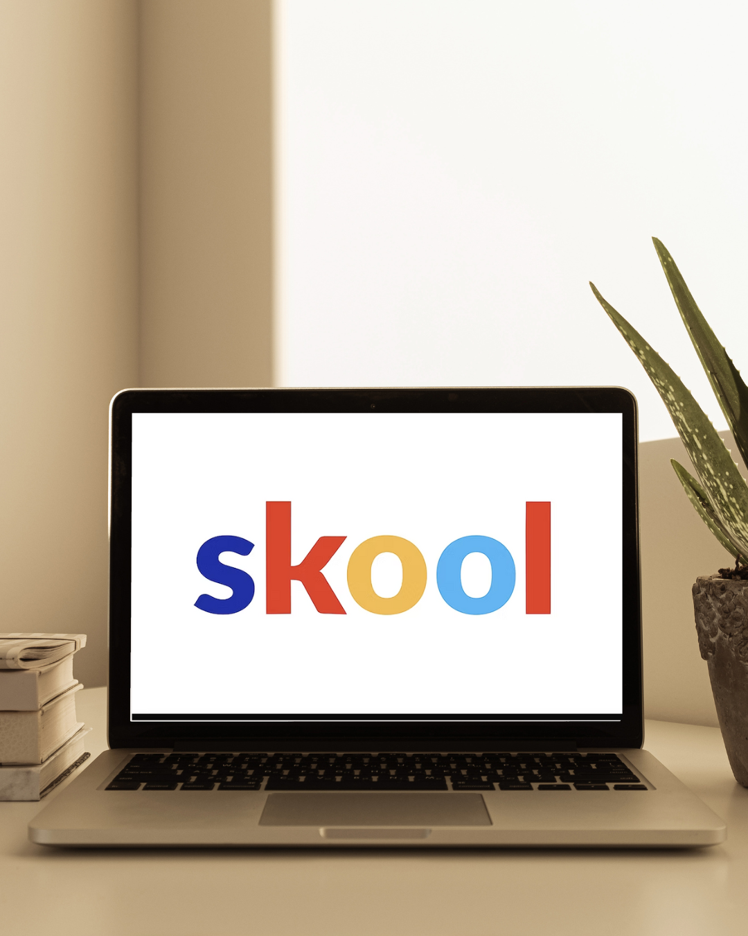 Laptop on a desk displaying the word 'skool' in colorful letters, with a plant on the right and stacked notebooks on the left.