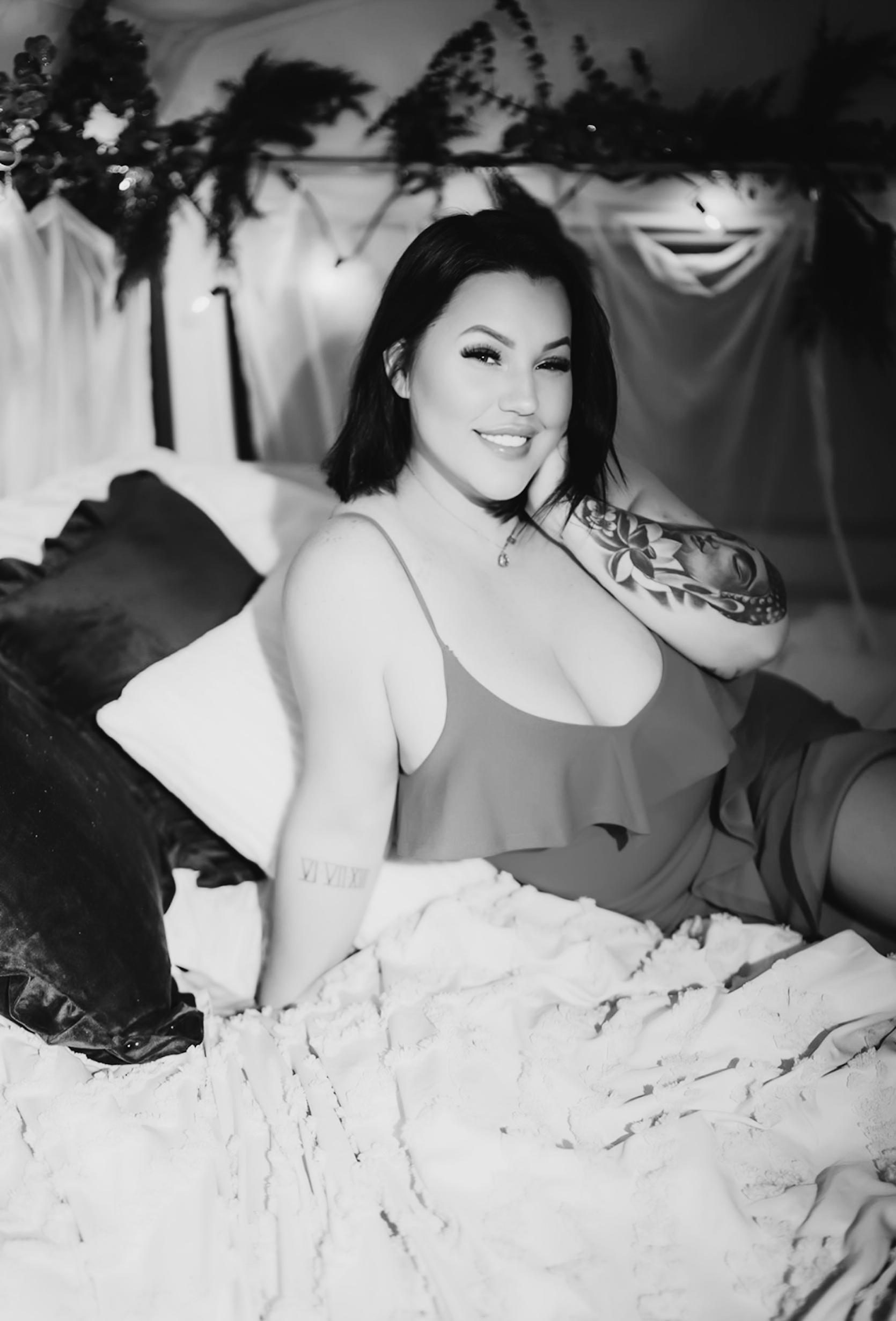 A woman with dark hair and tattoos smiling and winking while sitting on a bed with pillows, in a decorated room with hanging greenery and string lights.