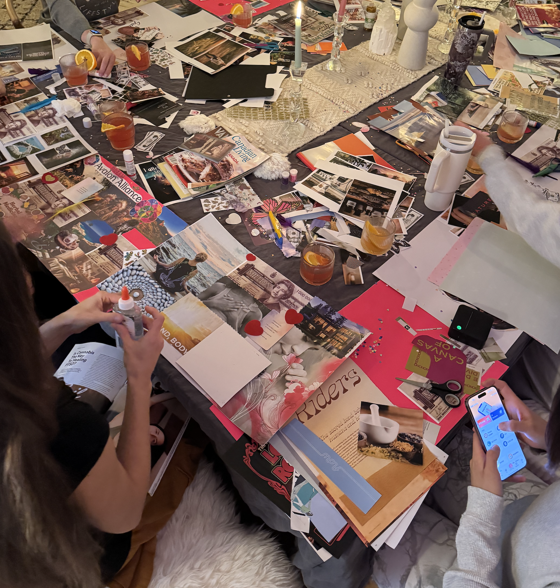 A cluttered table with various magazines, papers, scissors, glue, drinks, and two people; one is using a phone and the other is applying glue.