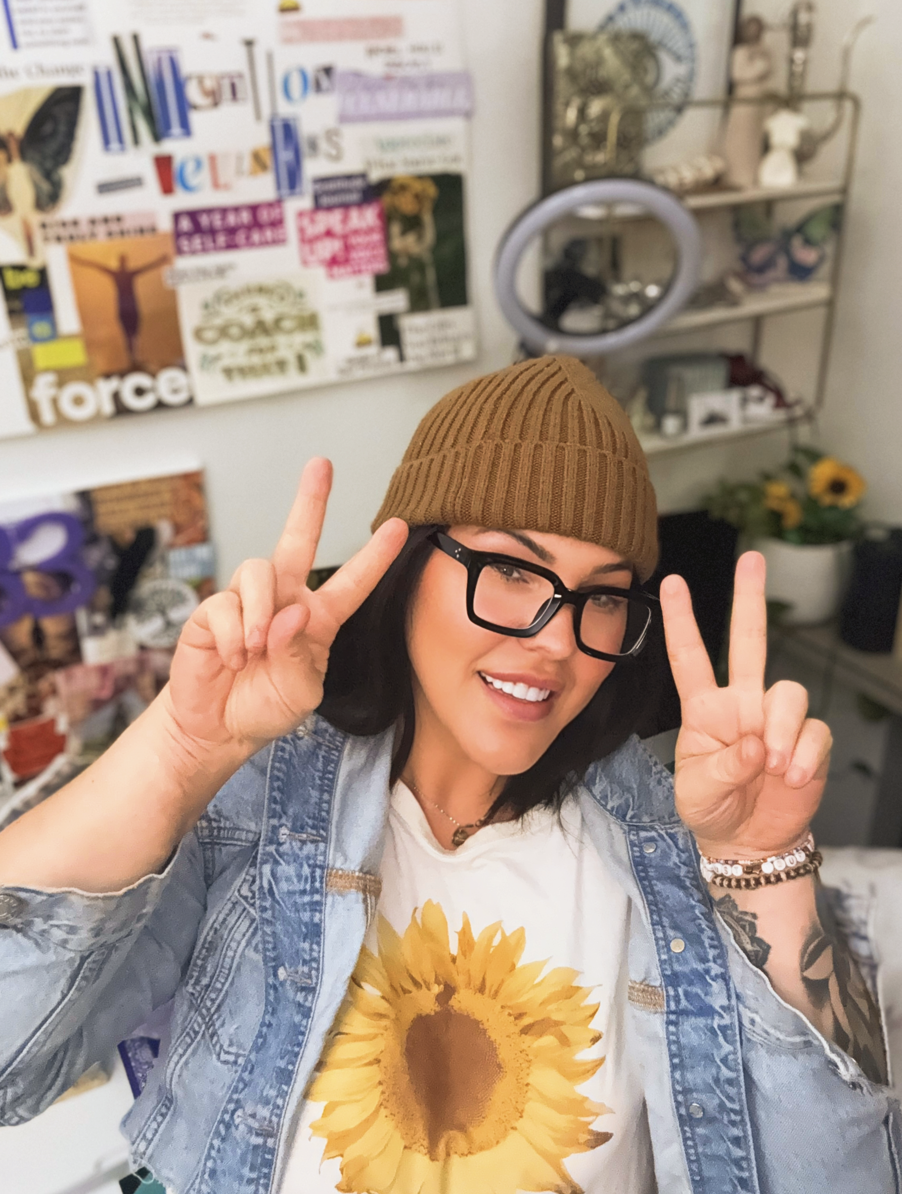 A smiling woman with black hair, wearing glasses, a brown knit beanie, a sunflower shirt, and a denim jacket, smiling and making peace signs with both hands in front of a cluttered room with posters, shelves, and a mirror.