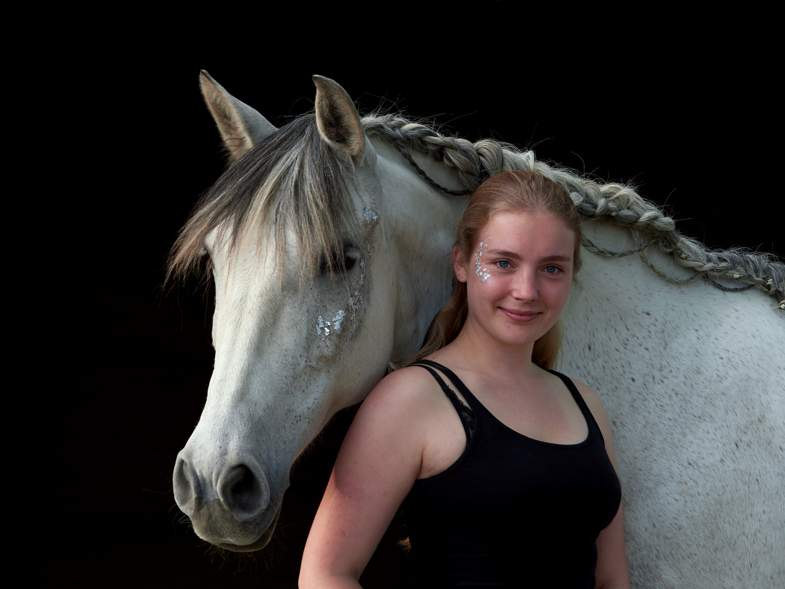Mona, ein Mädchen mit blondem Haar und silbernem Glitzer im Gesicht steht neben einem weißen Pferd mit geflochtener Mähne. Der Hintergrund ist schwarz.