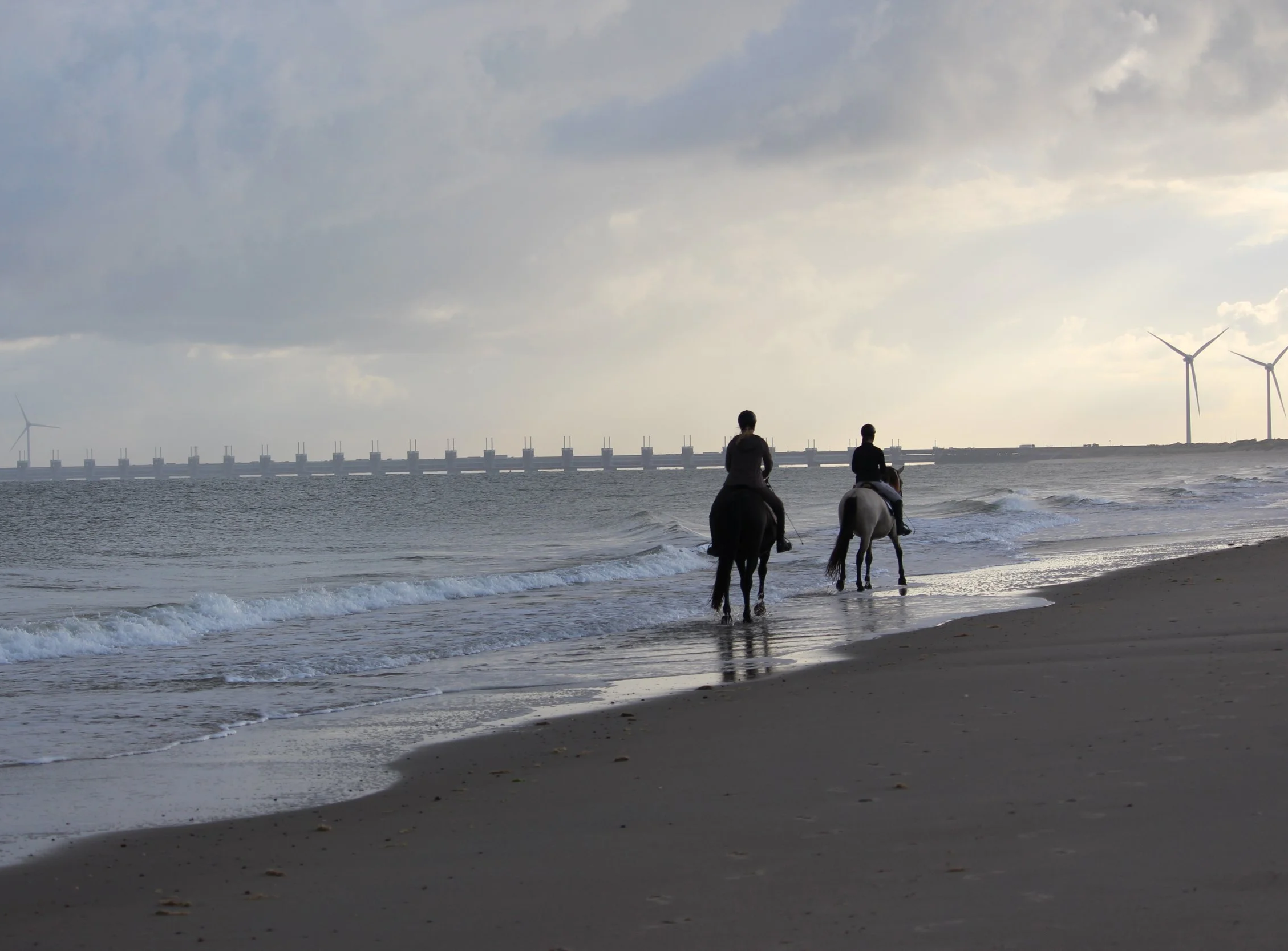 Zwei Personen reiten auf Pferden am Strand bei Sonnenuntergang, mit Windkraftanlagen im Hintergrund.