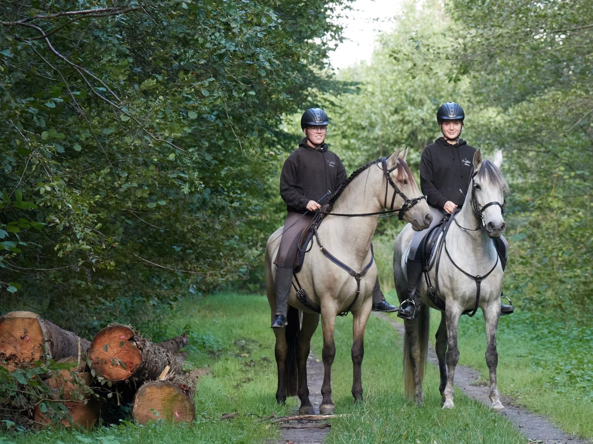 Zwei Frauen sitzen auf weißen Pferden in einem grünen Wald, tragen Helme und schwarze Hoodies, während sie auf einem schmalen Pfad reiten.