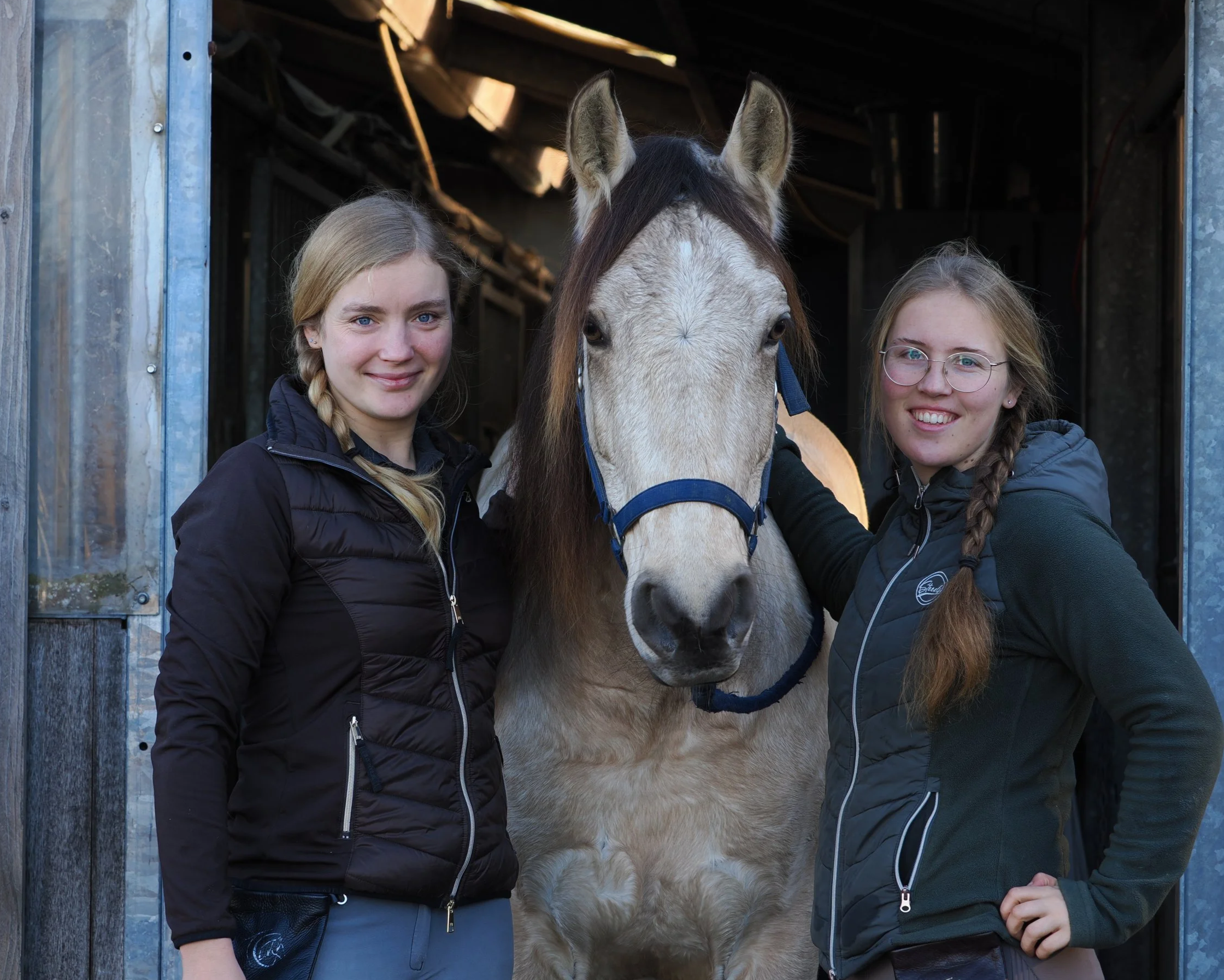 Zwei junge Frauen mit Zöpfen stehen neben einem weiß-braunen Pferd in einer Scheune, beide lächeln.