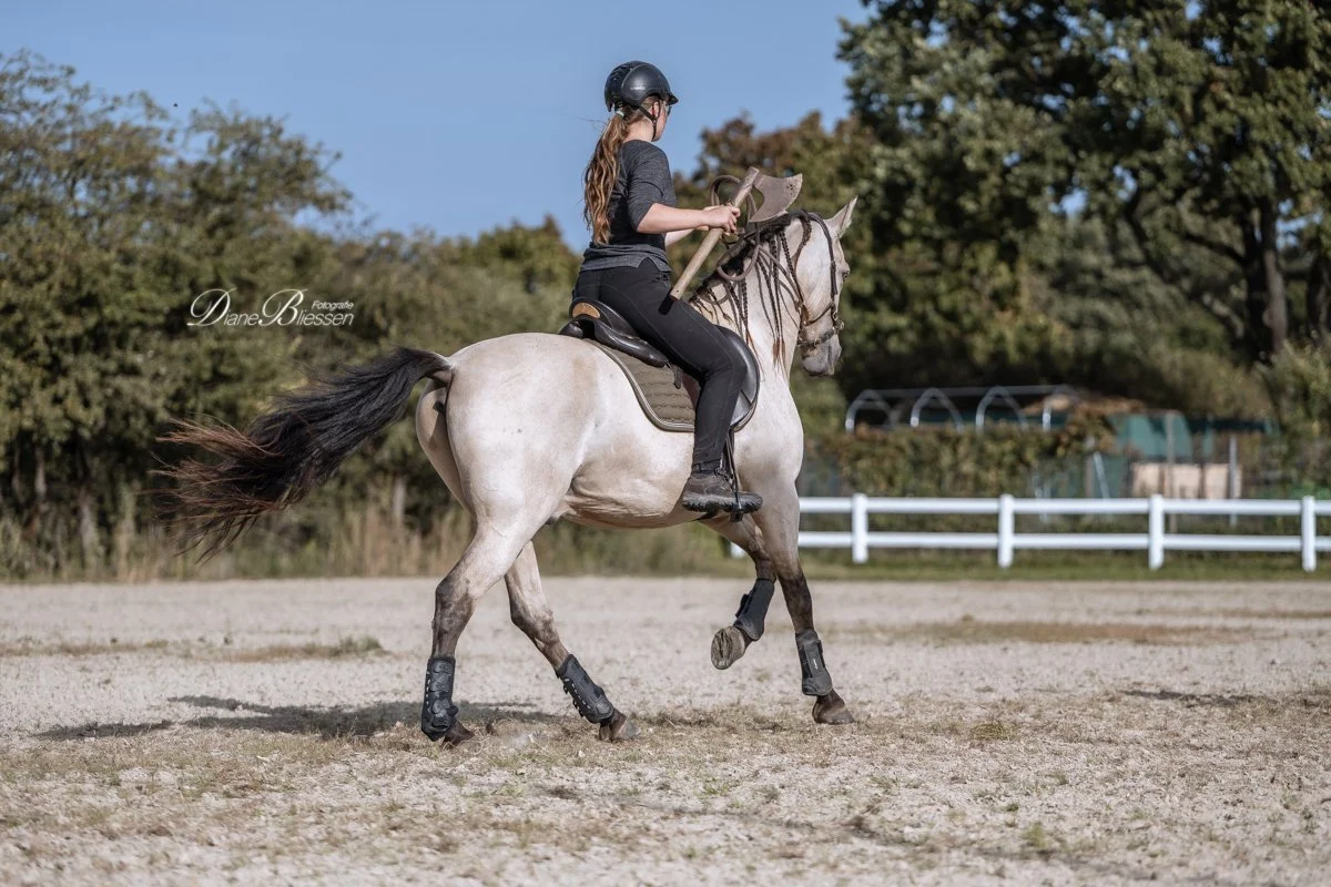 Mädchen reitet auf einem hellbraunen Pferd im Freien, trägt einen Helm und hält eine Axt in der Hand. Tano und Sarah beim Rittertraining