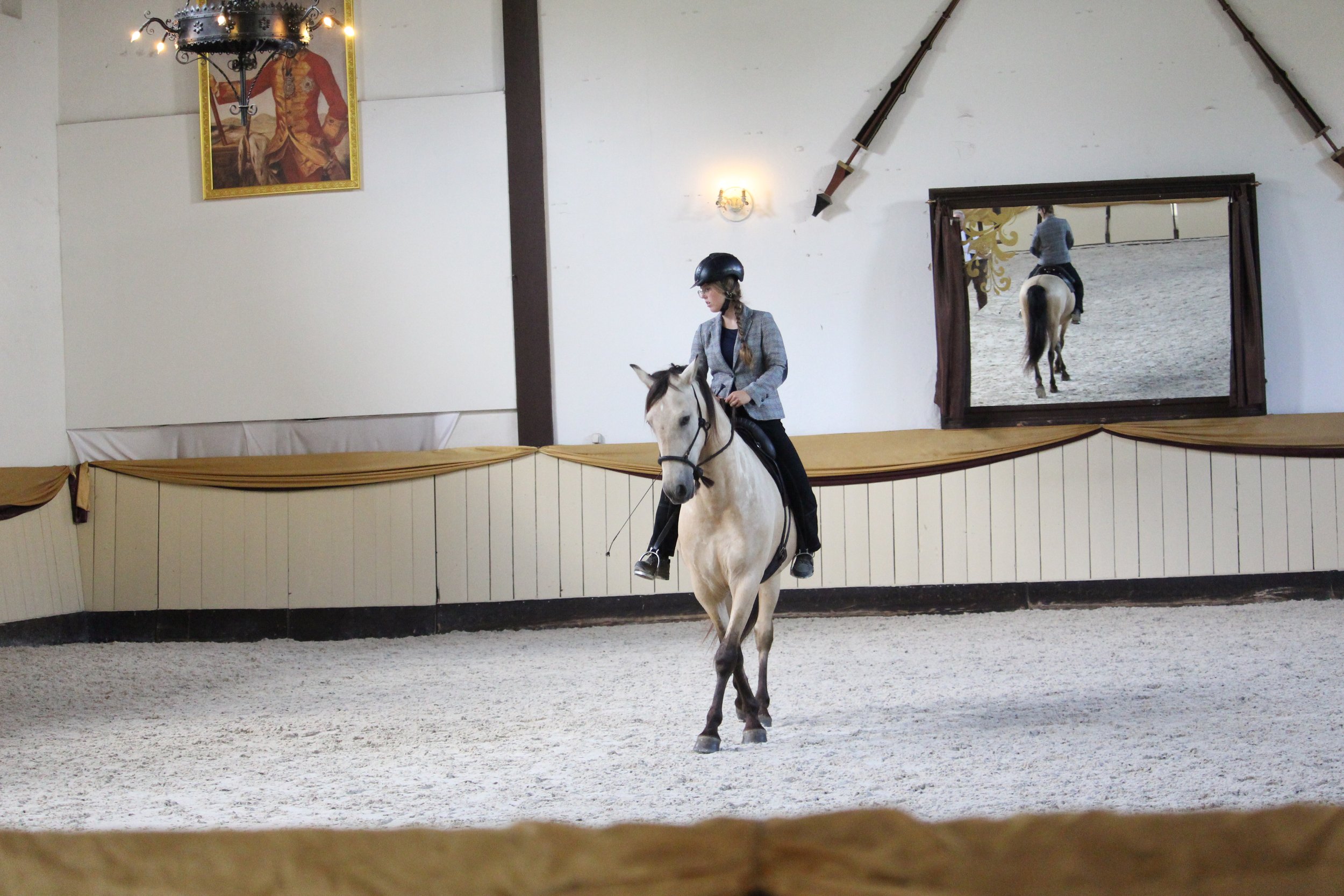 Eine Frau reitet auf einem hellbraunen Pferd in einer Reithalle. Im Hintergrund ist ein Spiegel, der die Reiterin zeigt. Das Bild zeigt Tano und Sarah in der Hofreitschule Bückeburg.