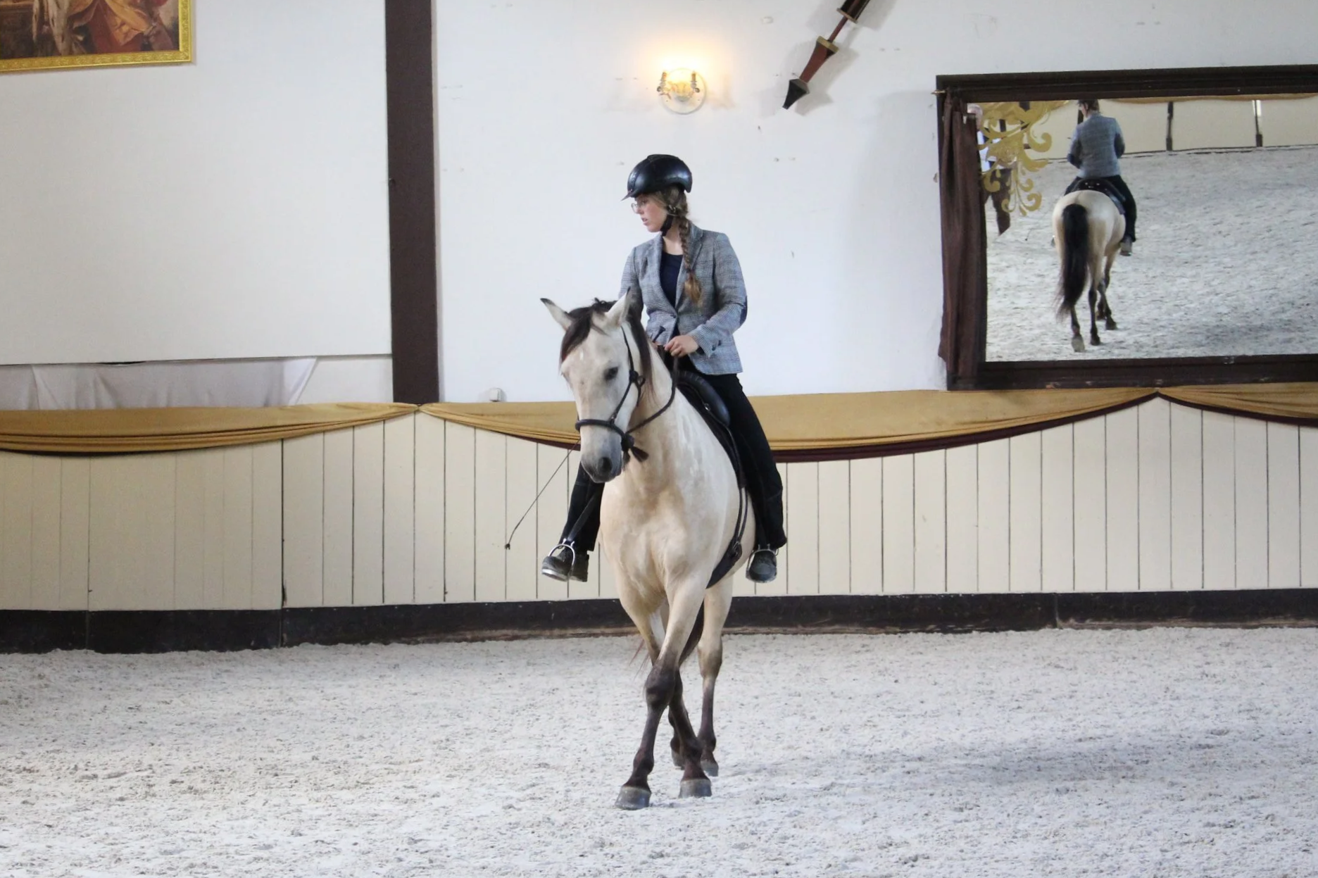 Junge Reiterin in Indoor-Reitbahn auf weißem Pferd, trägt Helm und Blazer, im Hintergrund Spiegel und Wandbild. Das Bild zeigt Sarah und Tano beim Training in der Hofreitschule Bückeburg