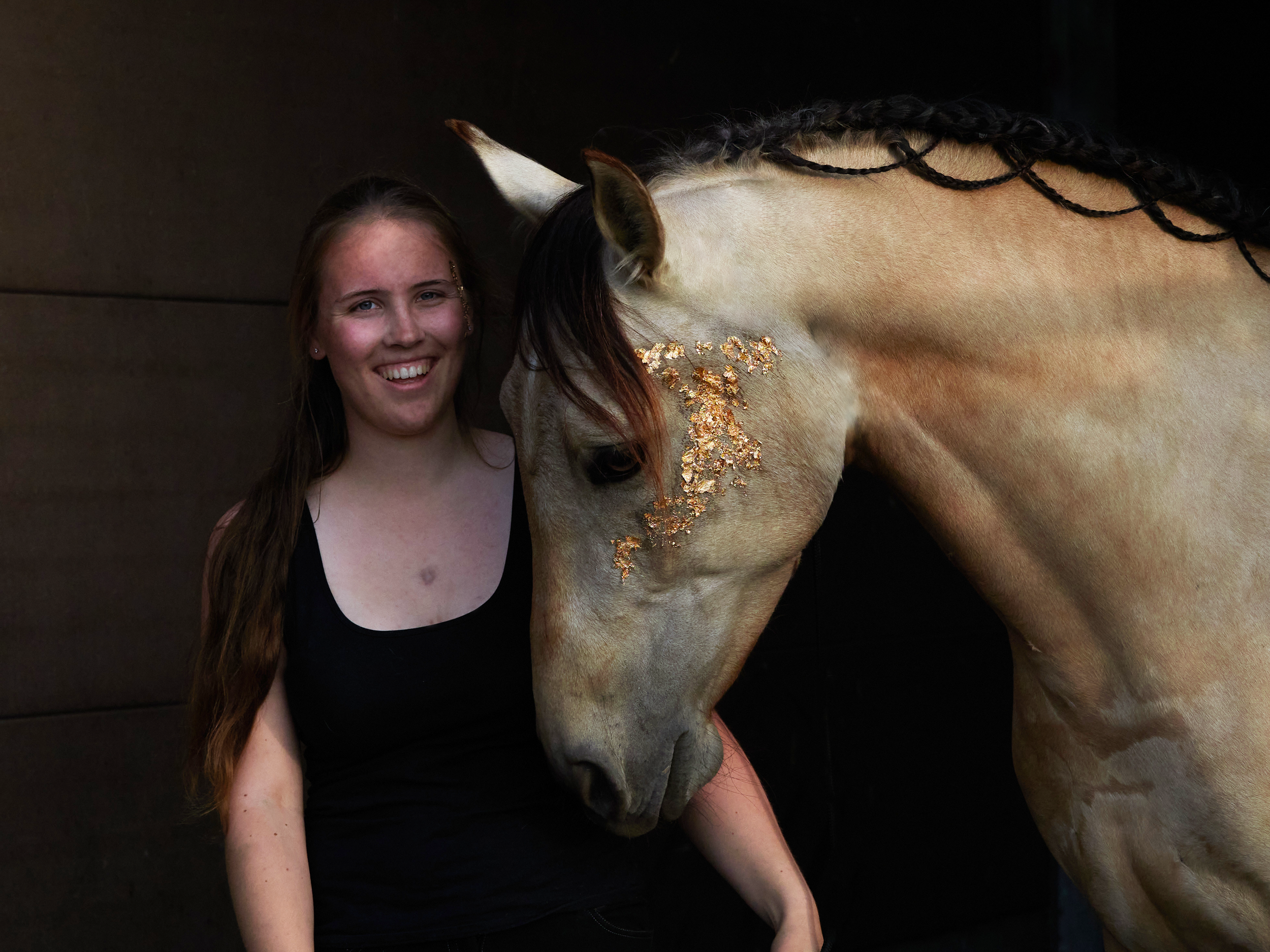 Sarah mit braunen Haaren und einem schwarzen Top neben Tano, einem goldenPferd mit geflochtener Mähne, das mit Goldflocken am Kopf dekoriert ist.