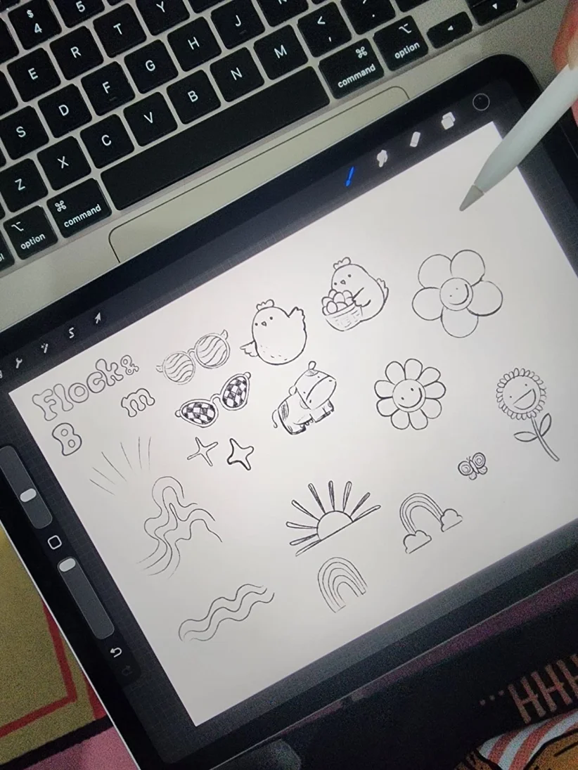 Digital drawing of various cartoon-style doodles including a chick, a flower, a rainbow, a sun, and other decorative elements on a tablet, with a stylus pen and a keyboard visible.