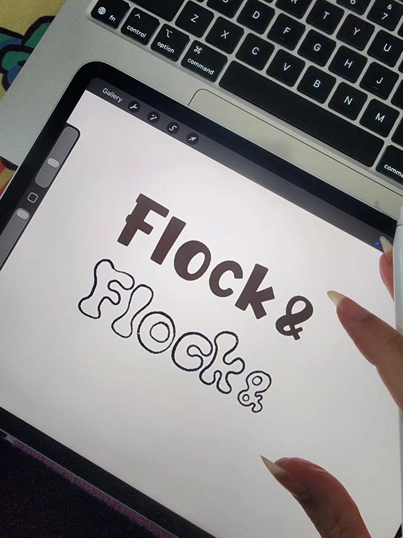 Tablet screen displaying the text 'FLOCK & FLASH' with the second word outlined in a bubbly font, held by a person's hand near a laptop keyboard.