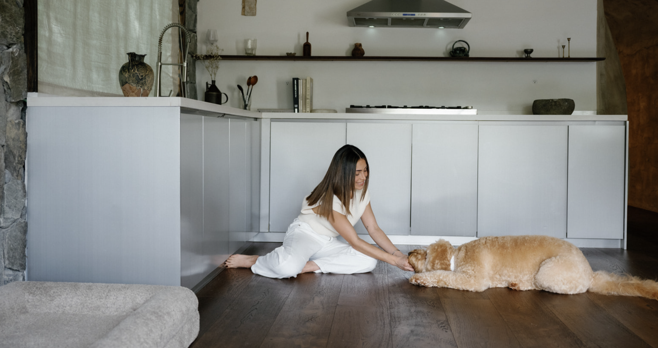 A woman in white pajamas playing with a golden retriever puppy on a hardwood floor in a modern kitchen.