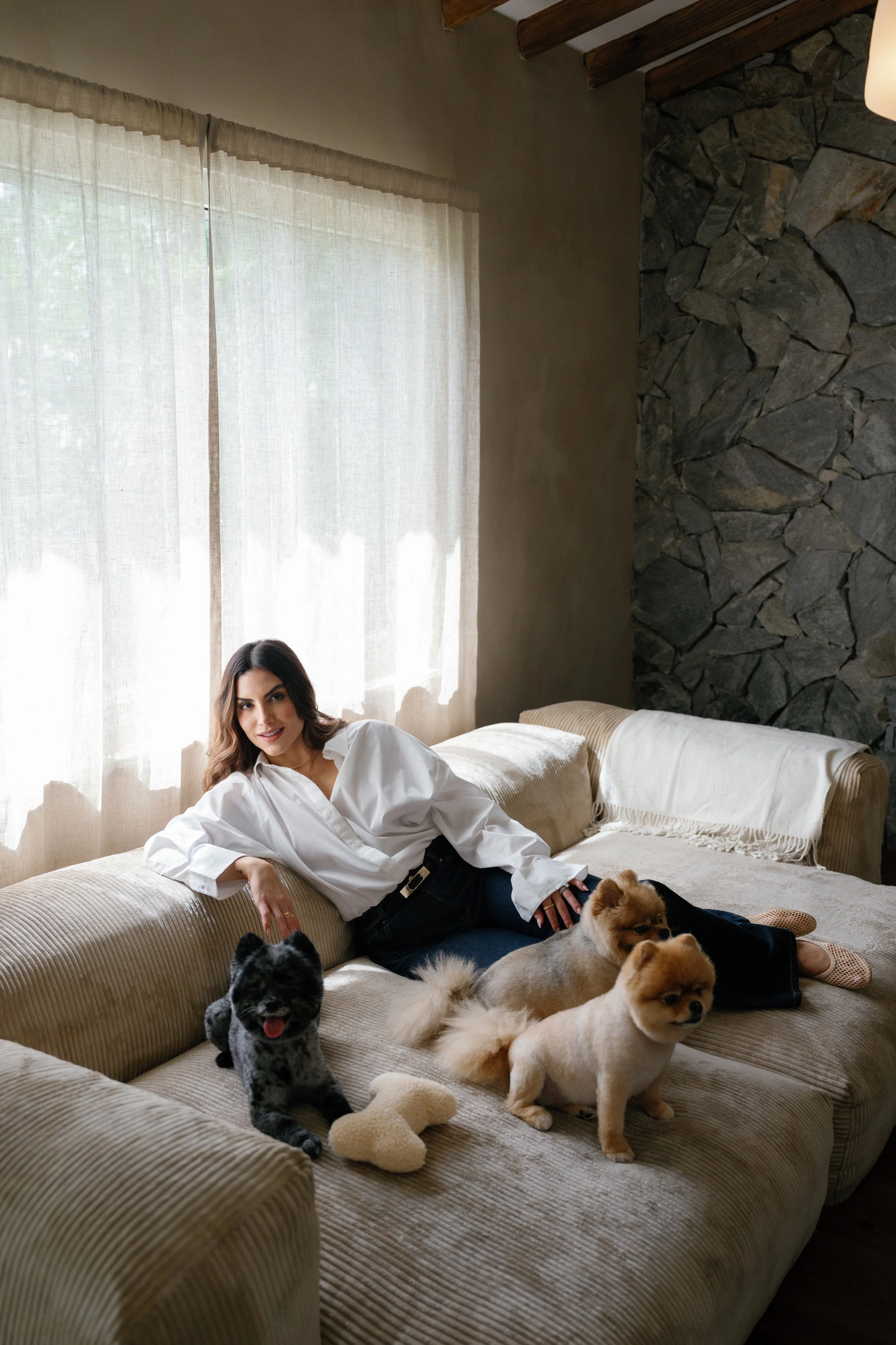 A woman relaxing on a beige couch with three small dogs in a cozy living room with a stone accent wall and a large window with curtains.