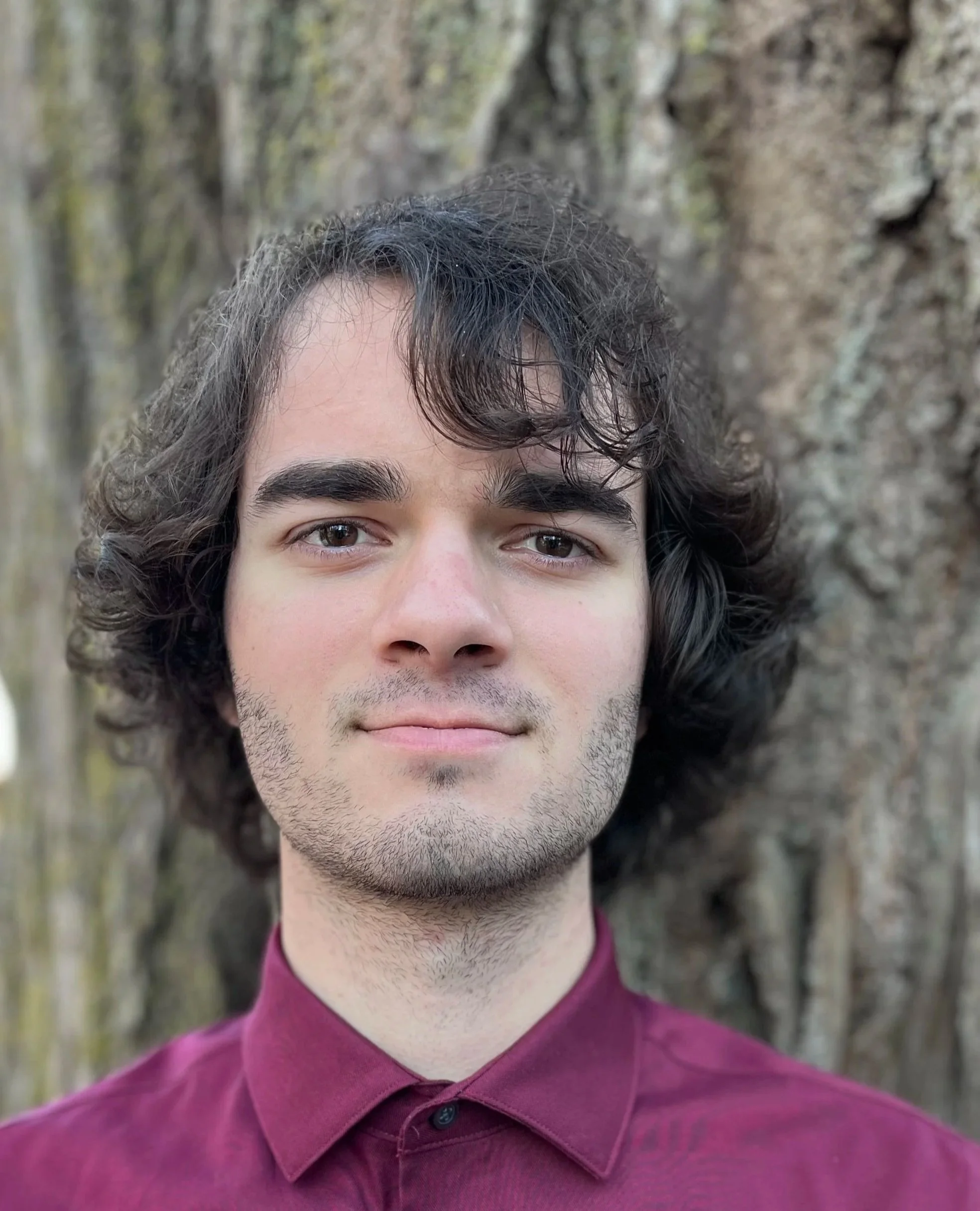 A photo of writer Tom Epic wearing a maroon collared shirt, standing outdoors in front of a tree.