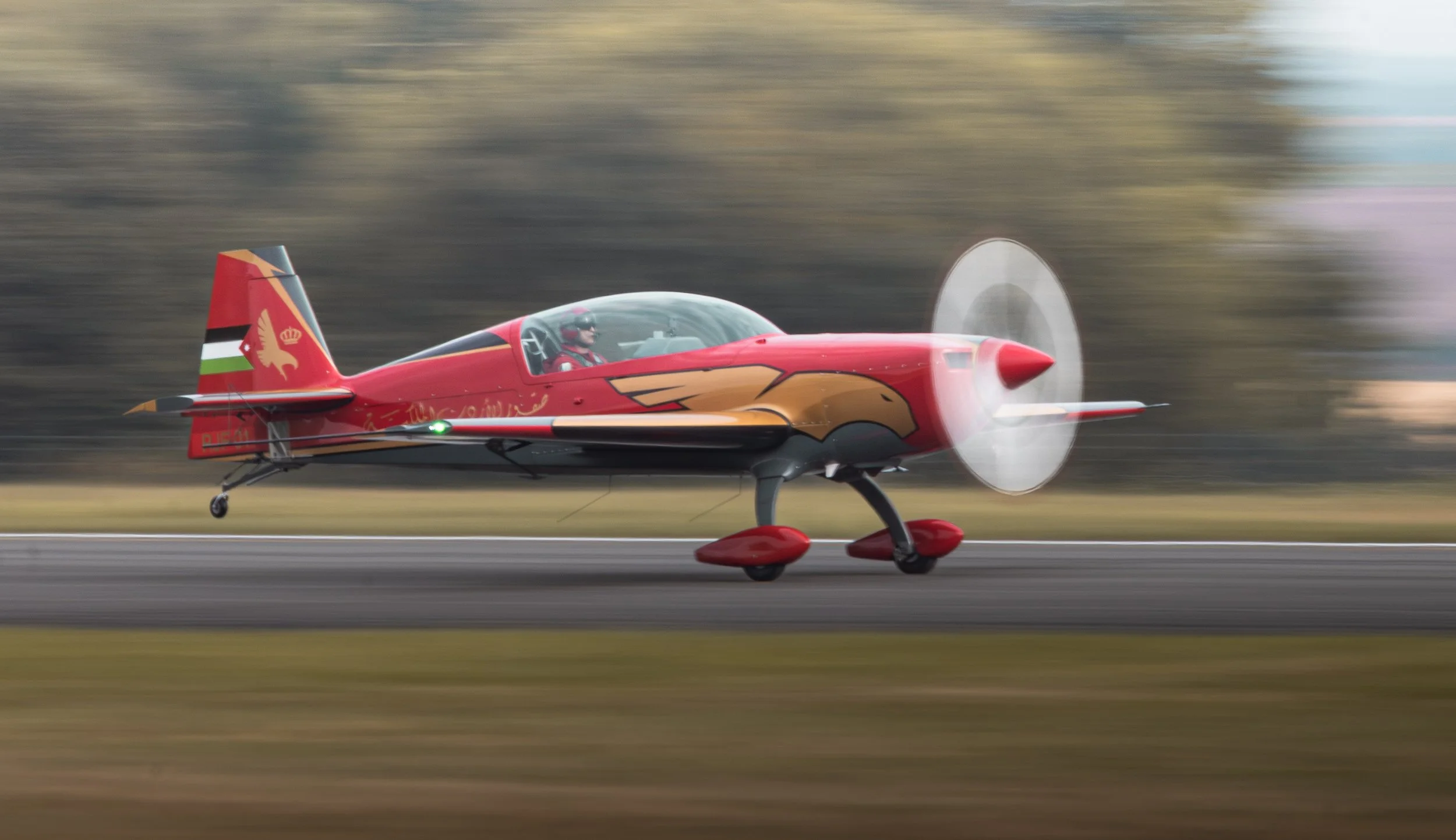 Avion de voltige rouge avec des décorations dorées, en mouvement rapide sur une piste, avec flou de fond, portant un pilote avec casque et visière.