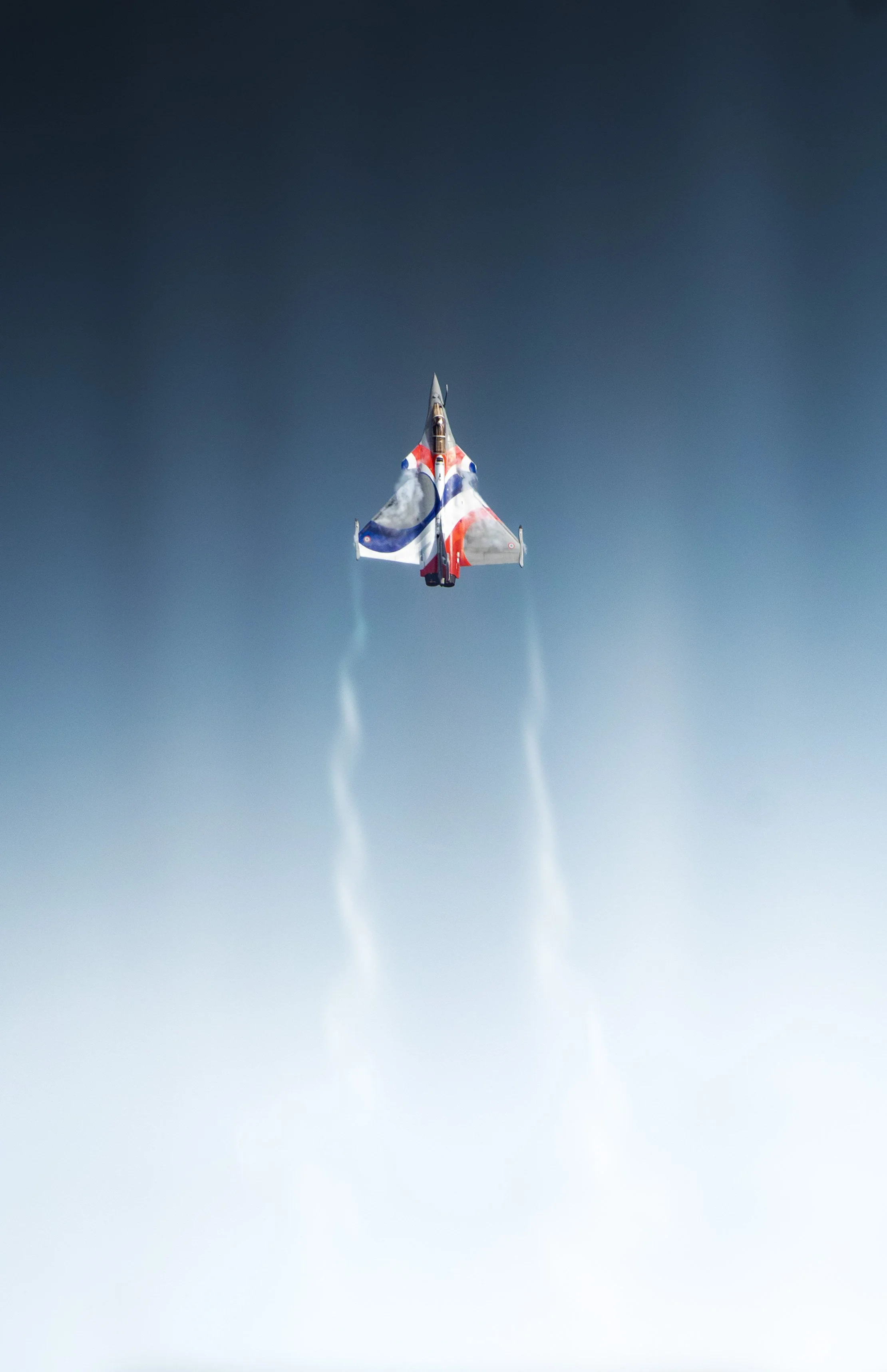 Un avion de chasse Français Rafale solo display RSD avec un camouflage aux couleurs du drapeau français, volant en altitude et laissant des traînées de condensation.