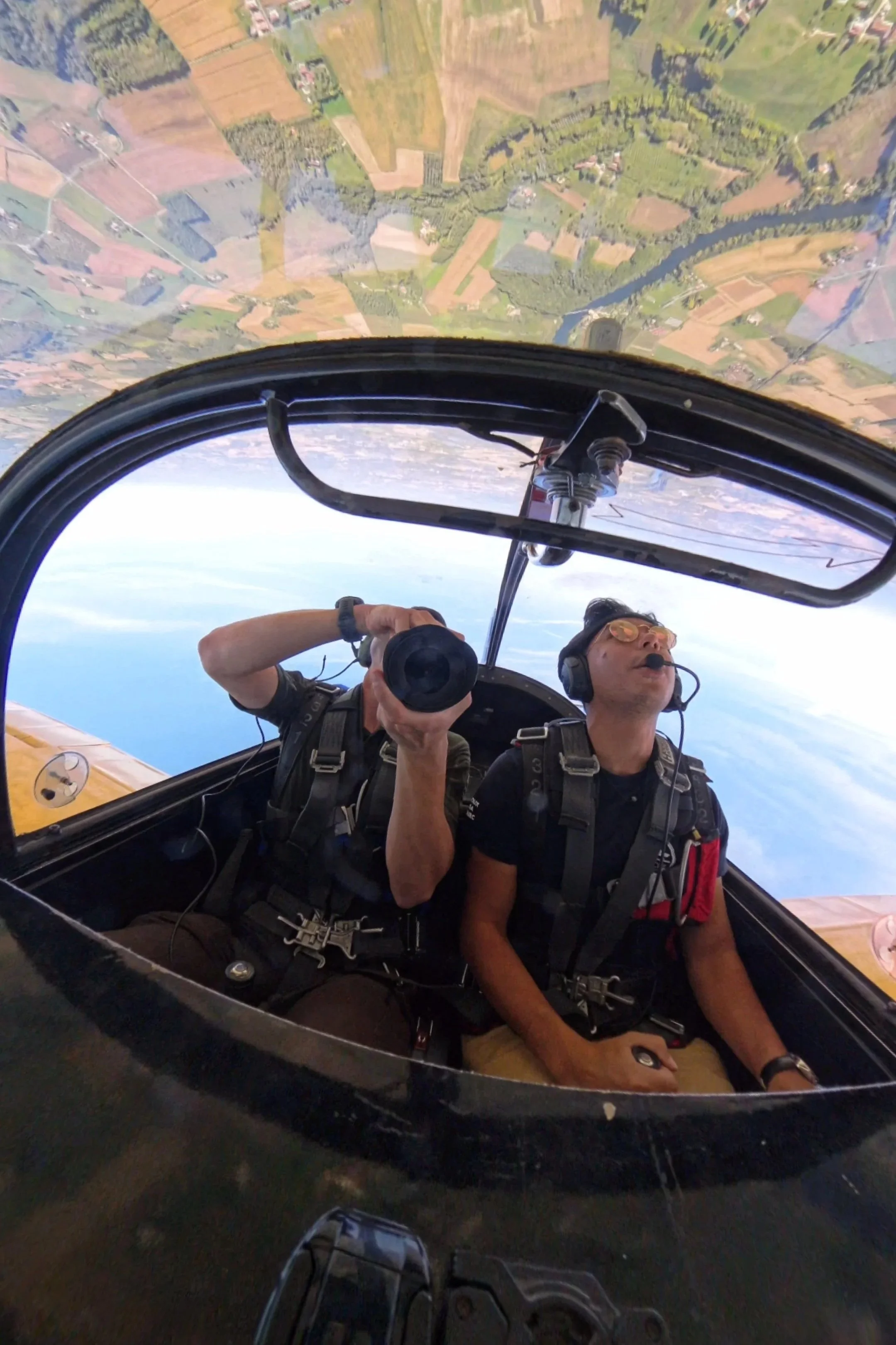 Deux pilotes de voltige dans un cockpit d'avion, volant à haute altitude avec un paysage de campagne en dessous.